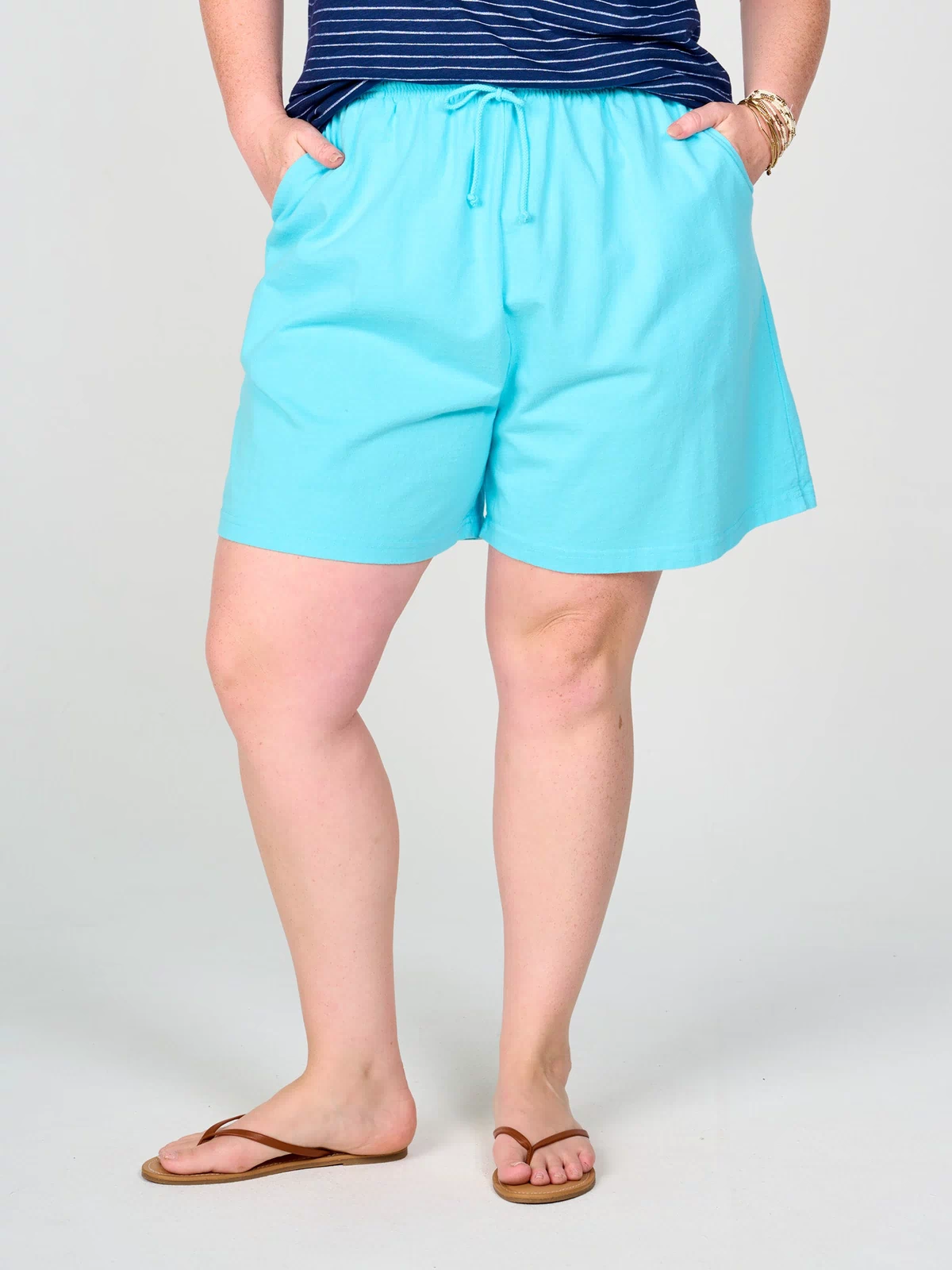 A person is shown from the waist down wearing Fresh Produce Clothes’ Plus Size Jersey Shorts in light blue with deep side pockets, styled with a navy striped shirt, brown flip-flops, and bracelets. The individual stands against a plain background with hands in the shorts’ pockets.
