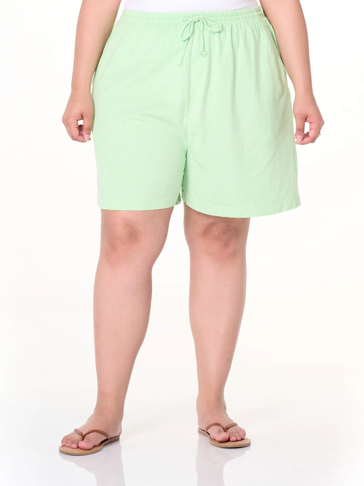A person stands against a plain white background, cropped at the shoulders, wearing Fresh Produce Clothes Jersey Shorts | Plus Size in light green with deep side pockets, paired with a white top and flip-flops.