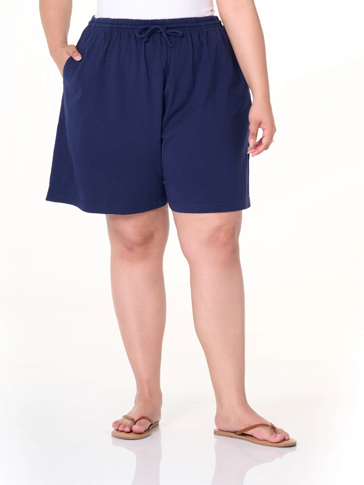 A person, with their head not visible in the image, is shown standing against a plain white background wearing a white top, tan flip flops, and navy blue Fresh Produce Clothes Jersey Shorts | Plus Size made from heavy cotton jersey with deep side pockets and a drawstring waist.