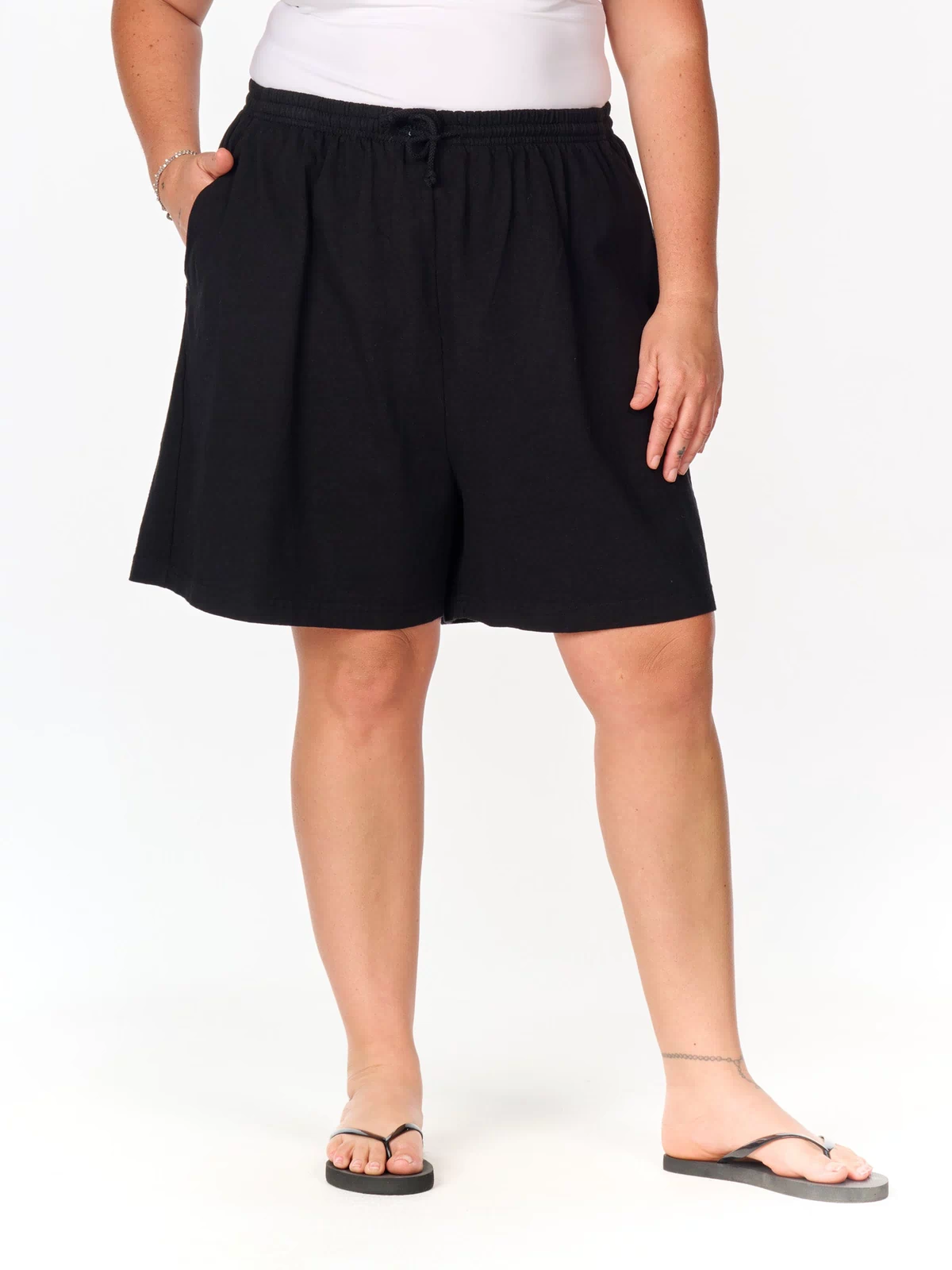 A person stands against a plain white background, one hand in their pocket, wearing Fresh Produce Clothes’ Jersey Shorts | Plus Size in black with deep side pockets, paired with a white top and black flip-flops.