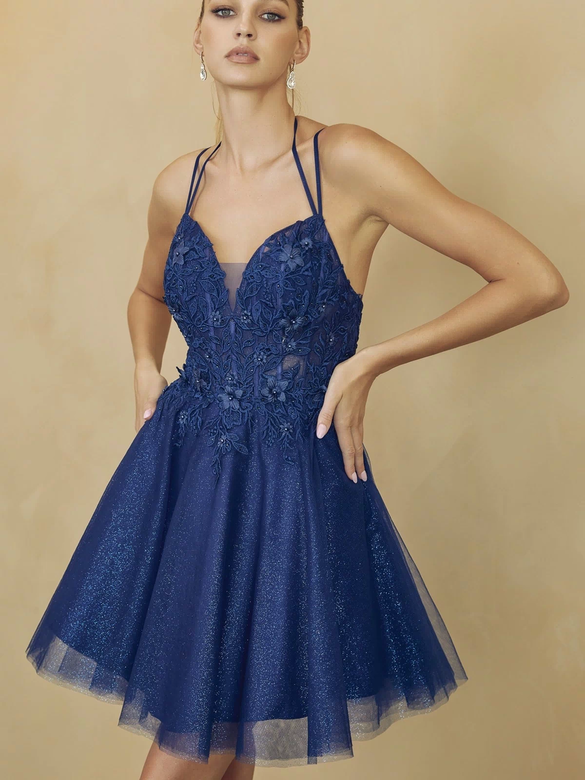A woman models the Glitter-Tulle Short A-Line Homecoming Dress 864 in navy blue, featuring floral embroidery and a glitter-tulle skirt, set against a beige background.