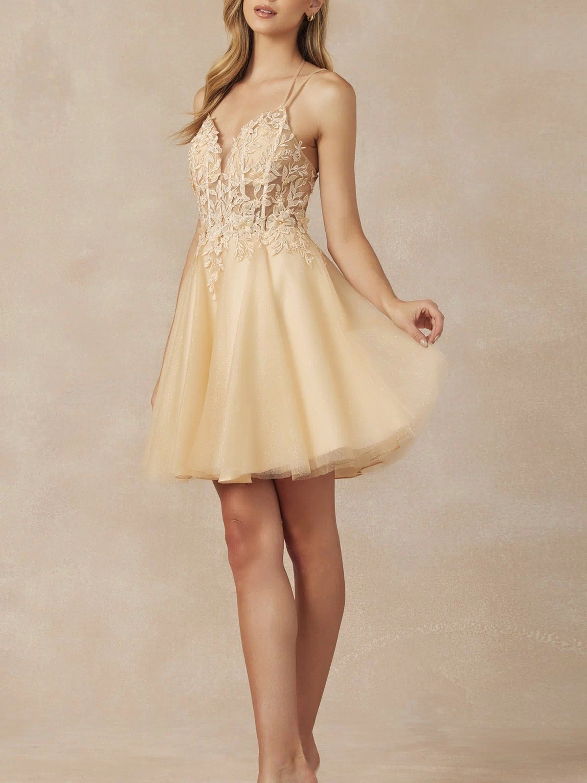 A woman wears the Glitter-Tulle Short A-Line Homecoming Dress 864, featuring spaghetti straps, a lace bodice, and a flared tulle skirt in light beige, standing before a neutral background.