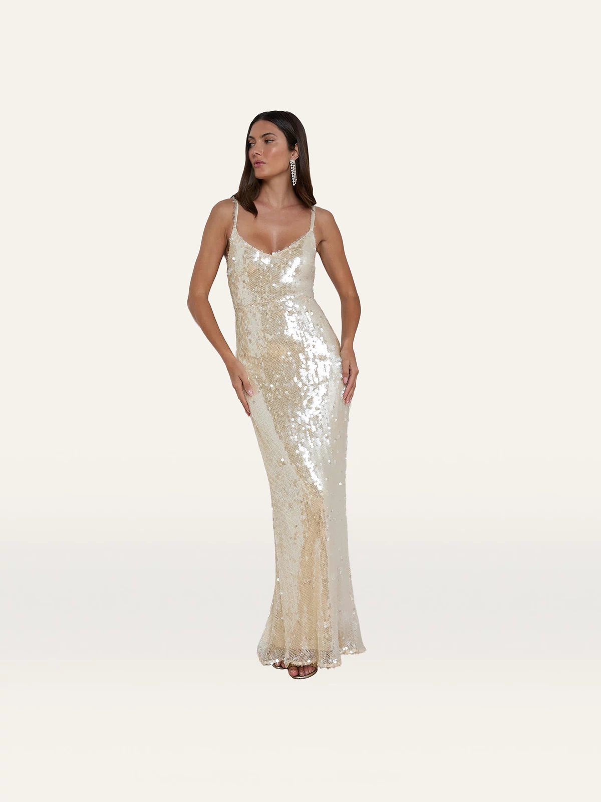 A woman wearing the Karma Sequin Maxi Dress—a sleeveless, floor-length gold dress featuring shimmering sequins and a V-neckline—stands against a plain white background. This elegant gown is perfect for NYFW 2026.