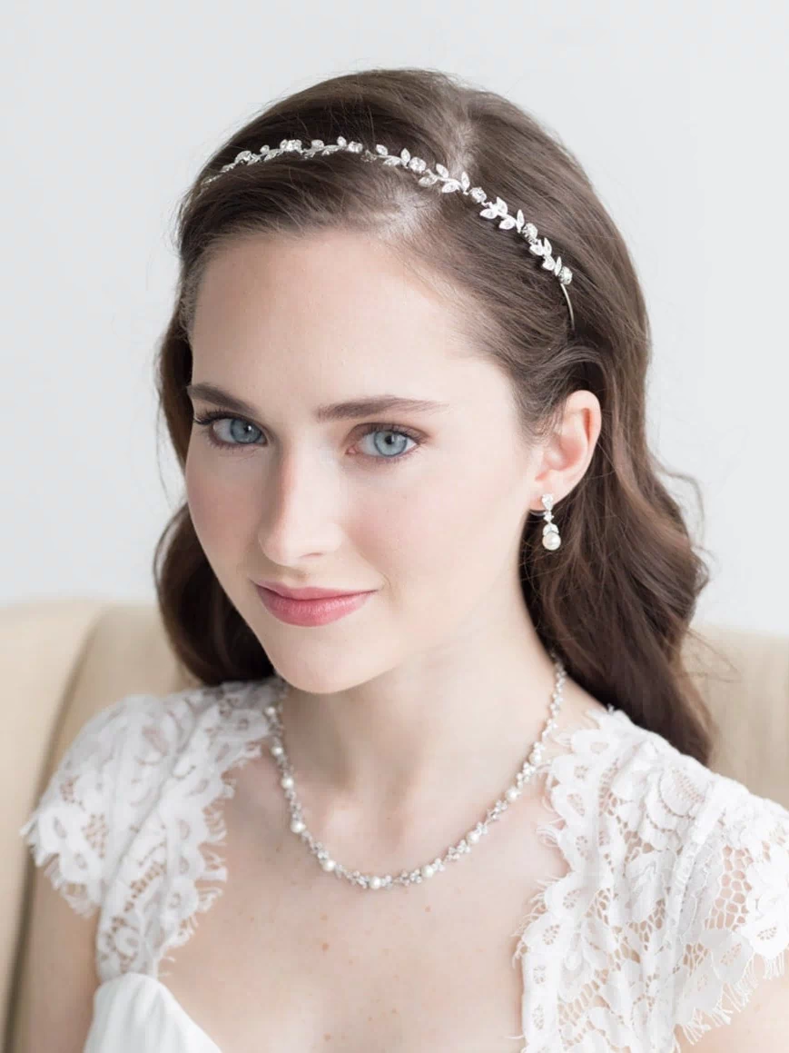A woman with long brown hair wearing a white lace dress and a headband with small white flowers, smiling at the camera.