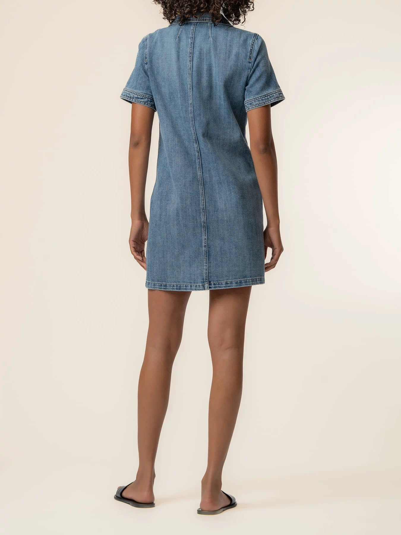 Woman wearing light blue denim dress with short sleeves and center seam, shown from back view.