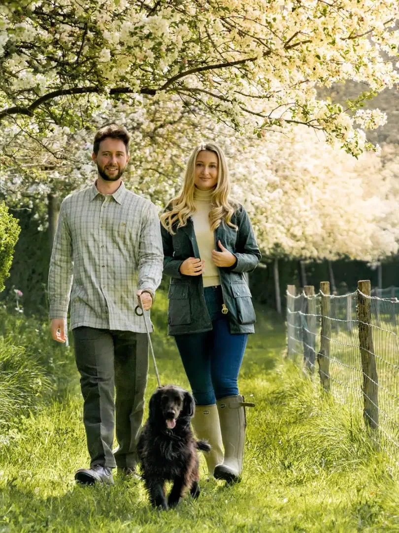 Couple in a traditional wax jacket walking a black dog under cherry blossoms