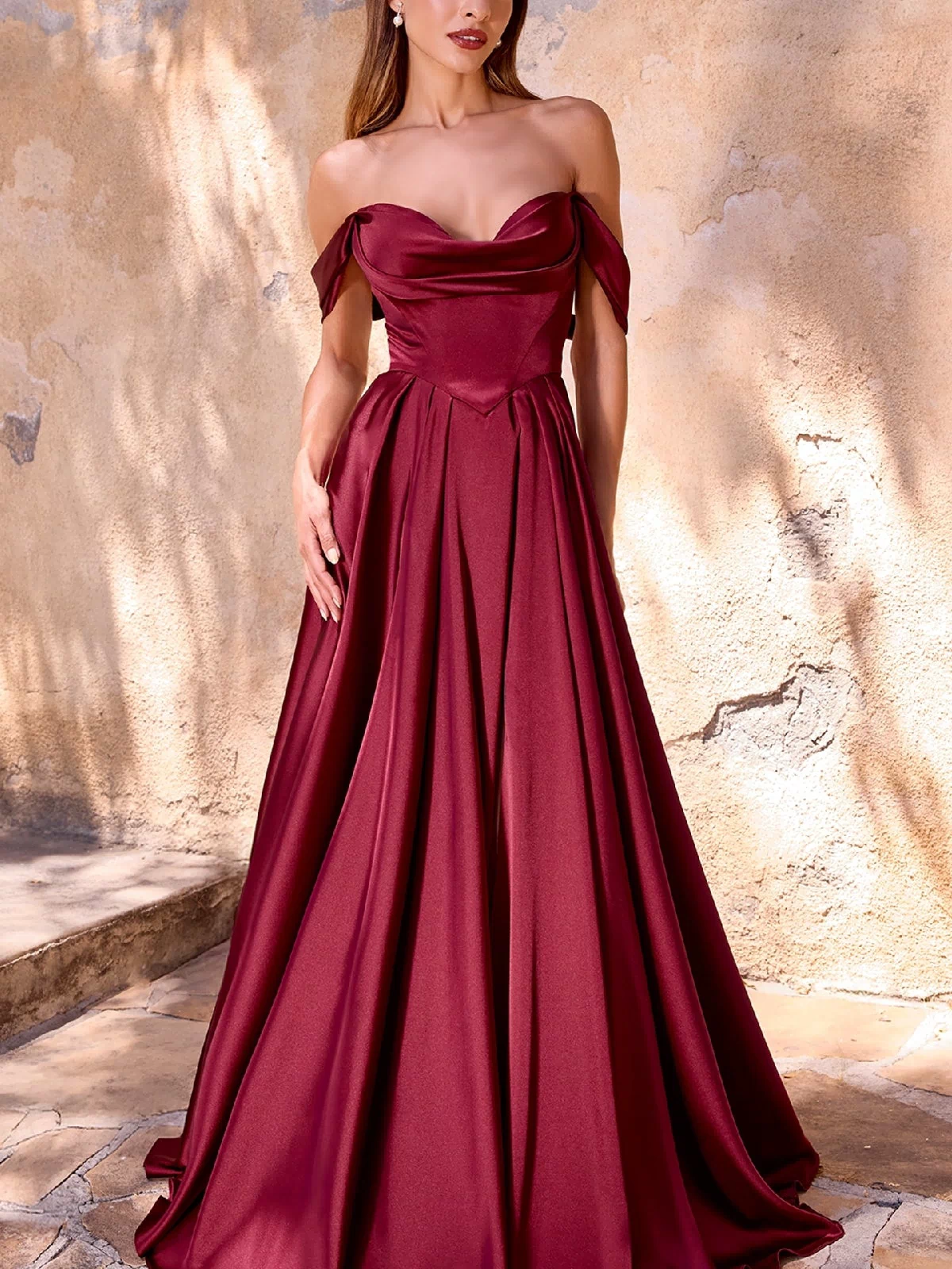 A woman stands before a textured beige wall, wearing the Ladivine 7449 Lace-Up Long Satin Prom Dress—an off-the-shoulder, floor-length burgundy gown with a full skirt.