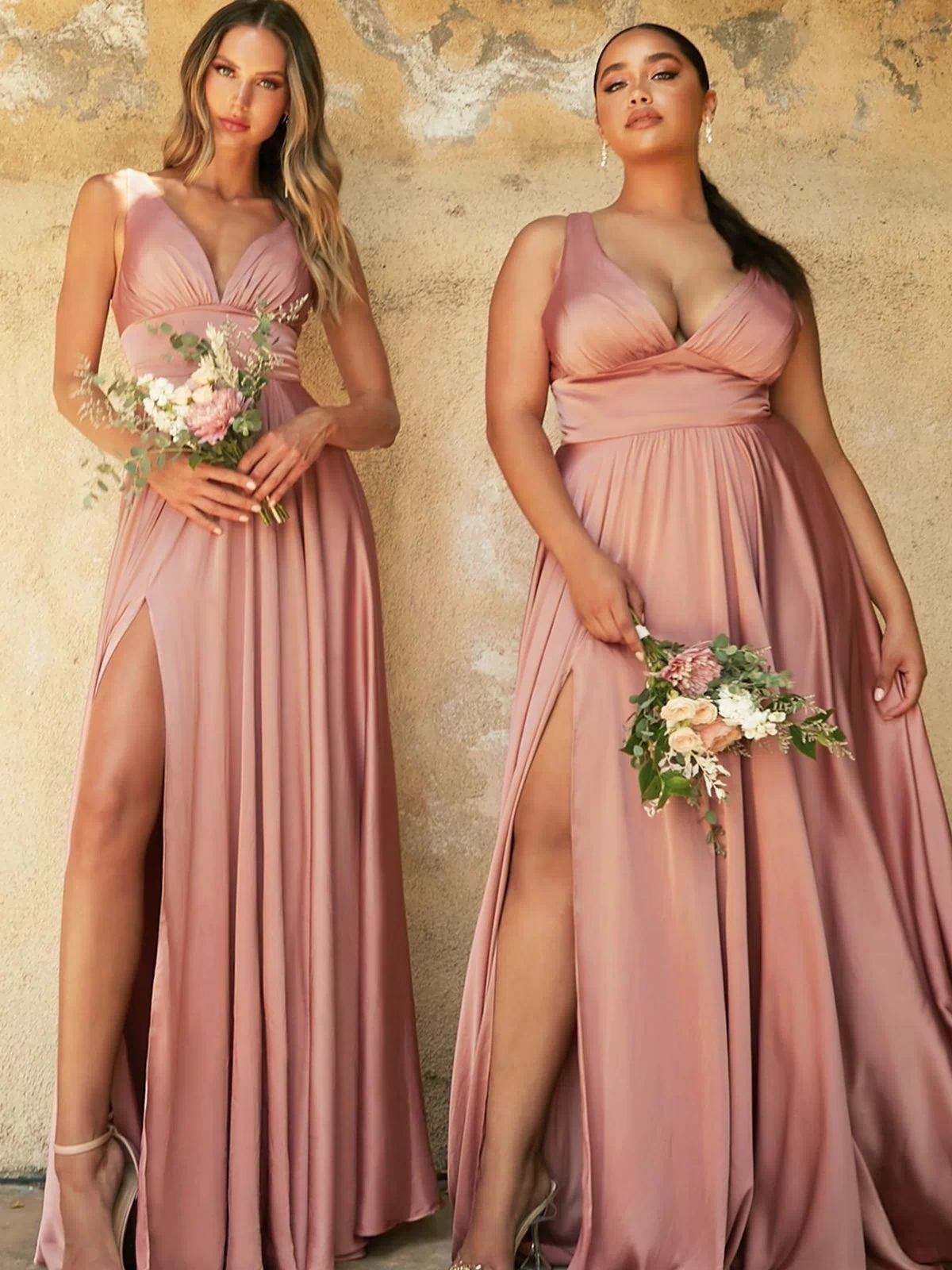 Two women in matching Empire-Waist Simple Satin Long A-Line Prom Dresses with thigh-high slits stand against a textured wall, each holding a small bouquet of flowers.