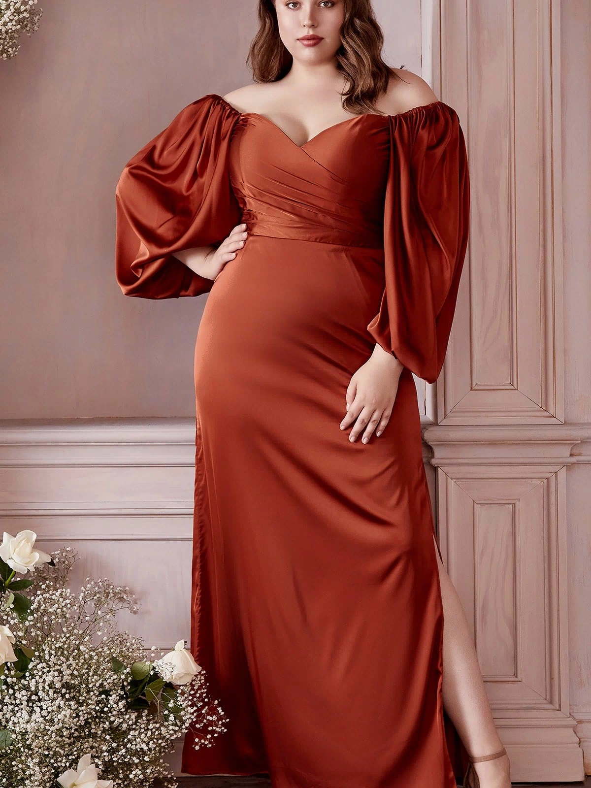 A woman in the Ladivine 7482C Plus Long Sleeve Long Prom Dress, featuring rust color and off-the-shoulder design, stands indoors by wooden paneling and white flowers.