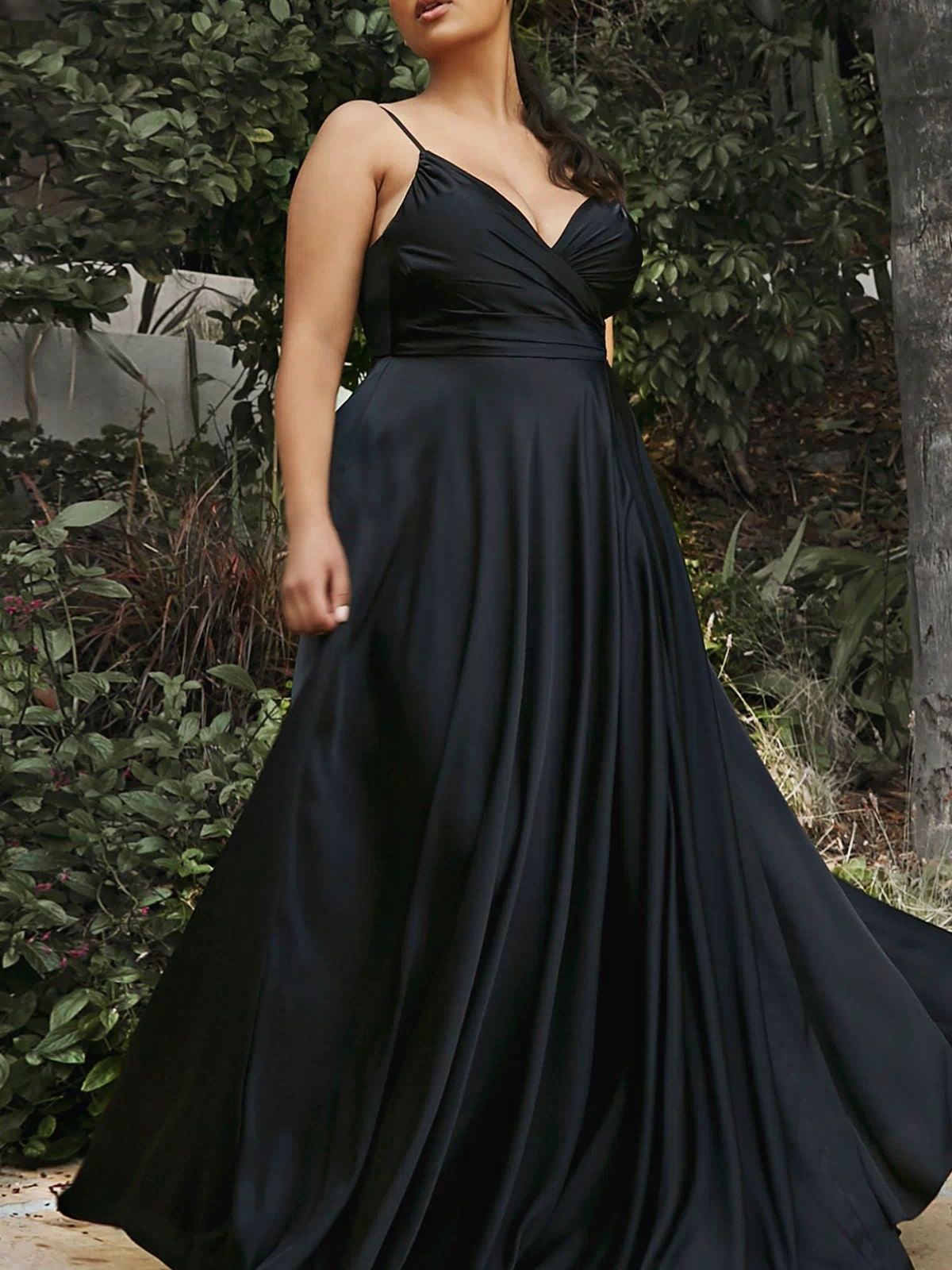A woman stands outdoors in a Classic Long Satin Formal Prom Dress: Ladivine 7485, a floor-length black gown with a deep V-neckline and A-line skirt, surrounded by greenery.