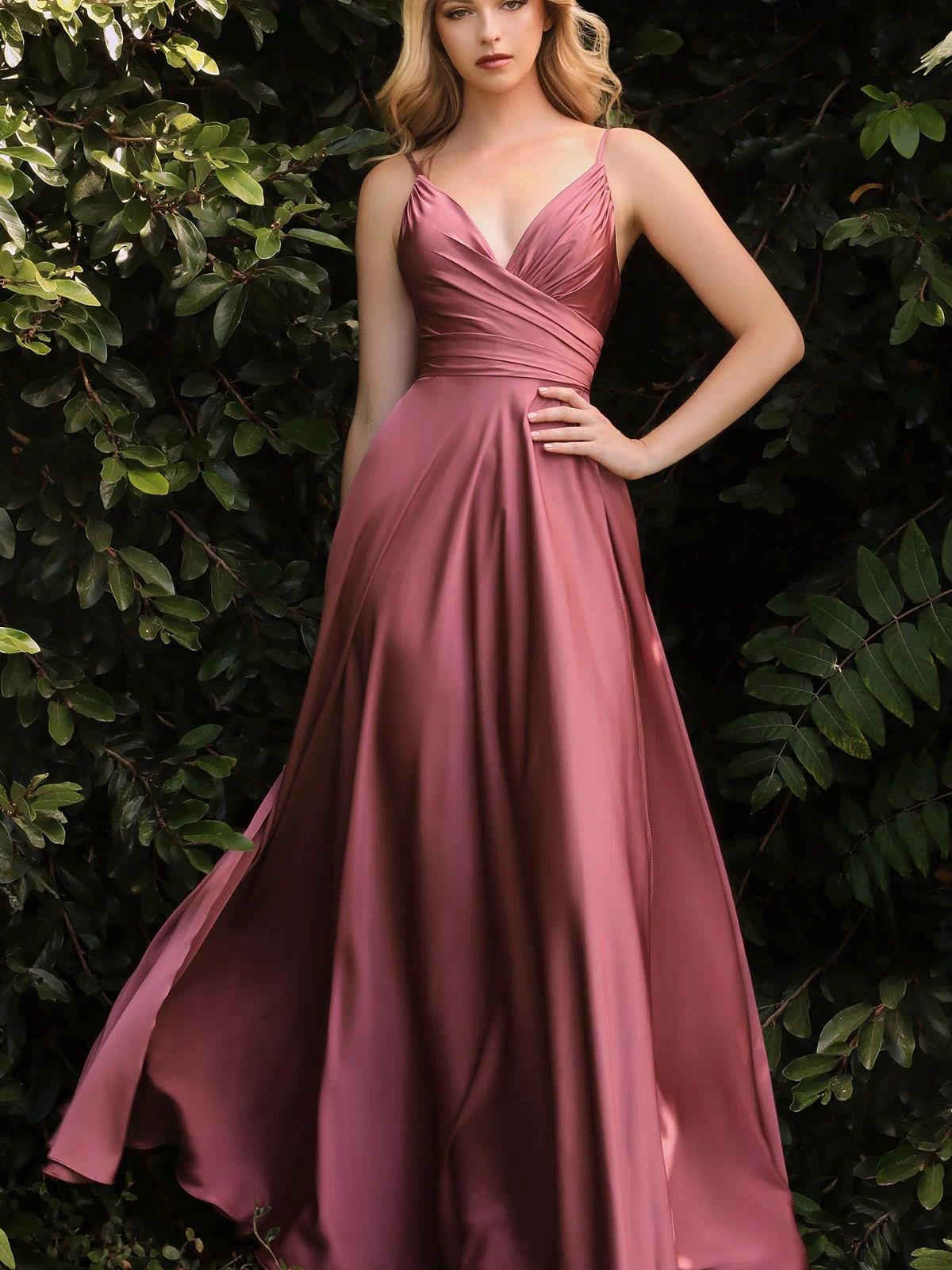 A woman stands outdoors before green foliage, wearing the Classic Long Satin Formal Prom Dress: Ladivine 7485—a floor-length, sleeveless rose satin gown with a V-neckline and pleated bodice.