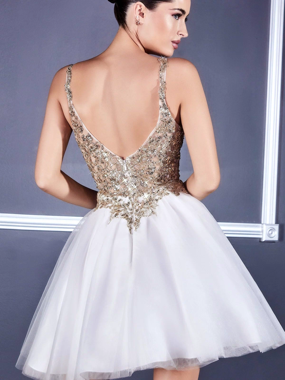 A woman stands facing away against a gray wall, wearing the Ballerina-Style Short Homecoming Dress 9239 with a gold sequin bodice, metallic-lace applique, and a short white tulle skirt.