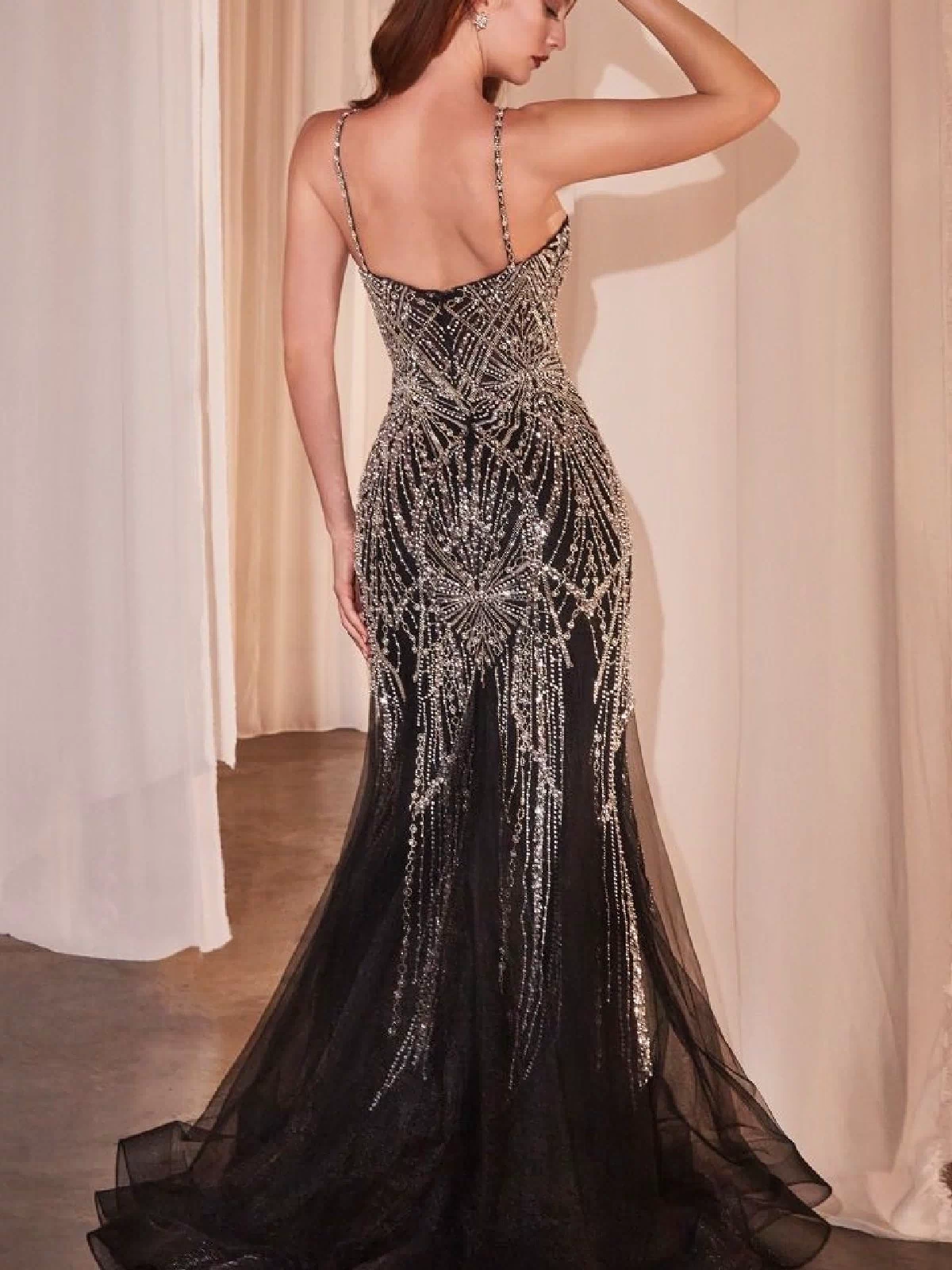 A woman poses with her back to the camera, wearing the Ladivine 9317 Long Black Beaded Prom Dress, featuring sheer accents and silver detailing, in a room with white curtains.