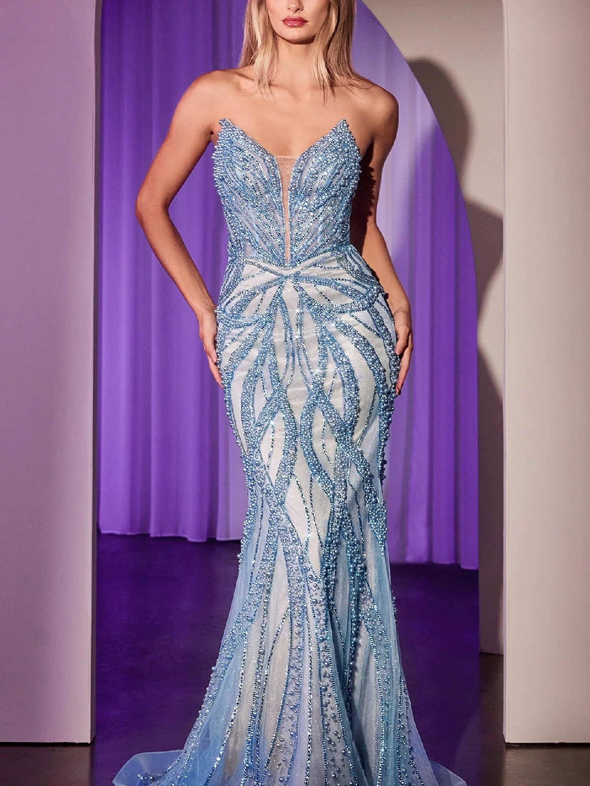 A woman stands in an arched doorway wearing the Ladivine CC0688, a strapless long beaded prom dress in blue with a fitted silhouette, wave-like patterns, and a flattering corset bodice.