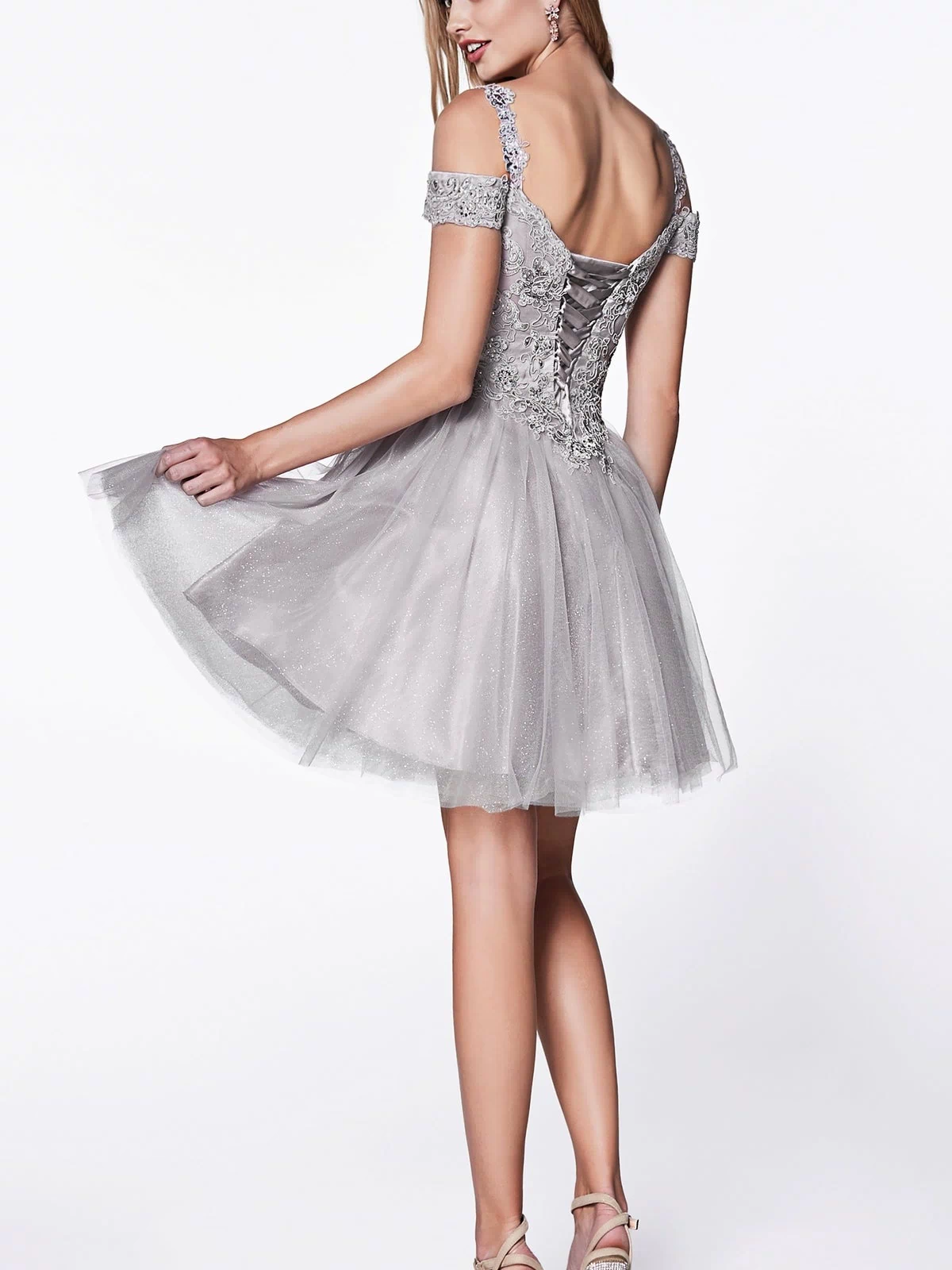 A woman poses in the Cold-Shoulder Short Glitter Homecoming Dress CD0132, a silver off-shoulder party dress with a lace-up back and tulle skirt, holding the skirt and finishing her look with high-heeled sandals.
