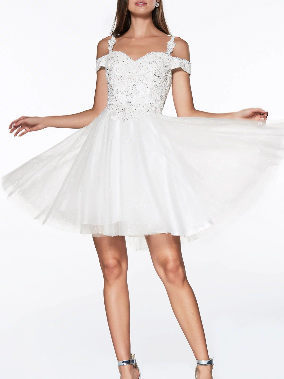 A woman models the Cold-Shoulder Short Glitter Homecoming Dress CD0132, featuring off-the-shoulder lace and sequin details, paired with silver high-heeled sandals. She holds out the flared sides to display its sparkling elegance.