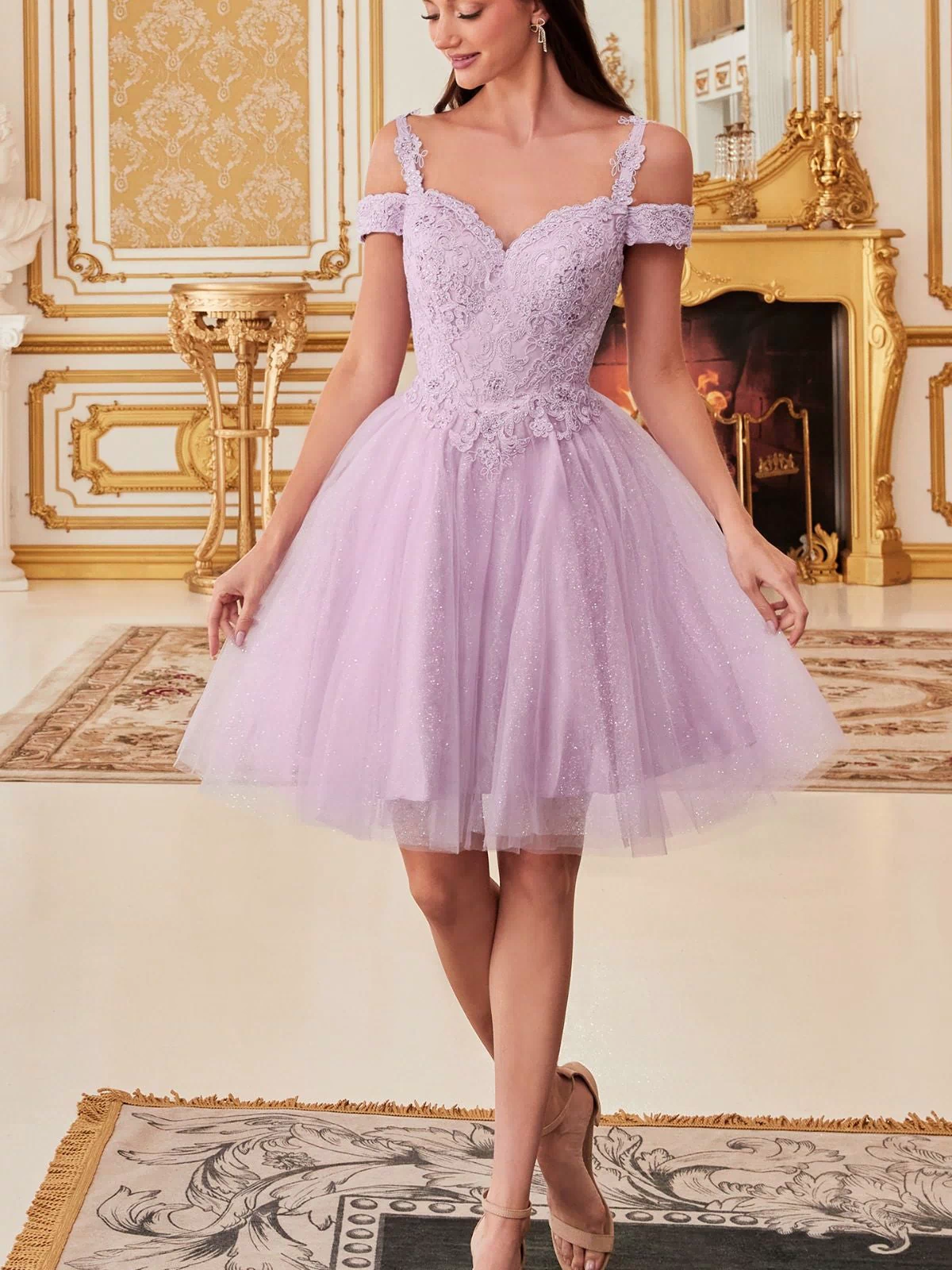 A woman wears the Cold-Shoulder Short Glitter Homecoming Dress CD0132, with lilac lace and tulle, standing in an ornate gold-accented room with a fireplace and chandelier, smiling as she looks to the side.