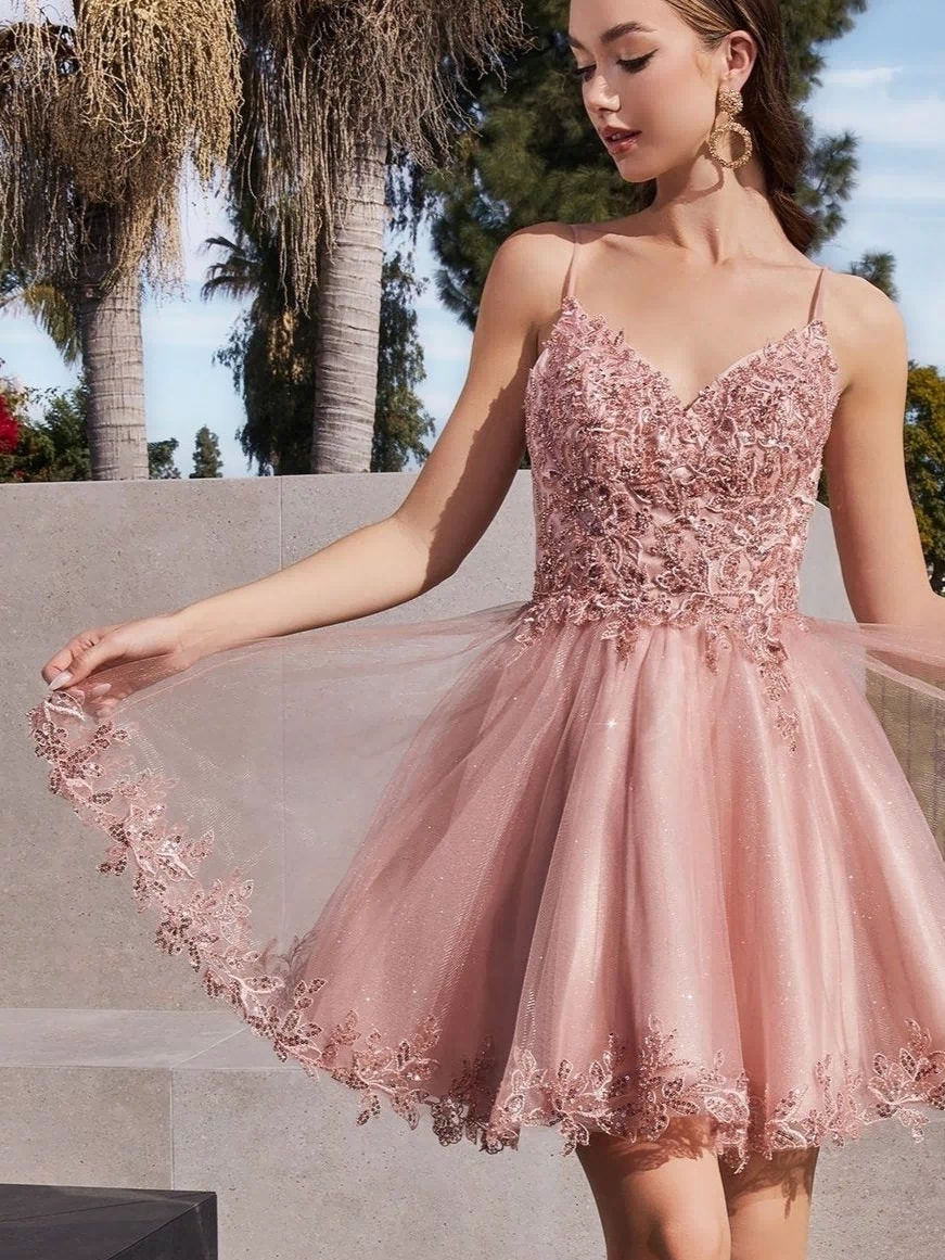 A young woman stands outdoors in the Short Homecoming Dress CD0213 with embroidery, holding the side of her pink, lace-embellished sleeveless skirt, with trees and concrete structures in the background.