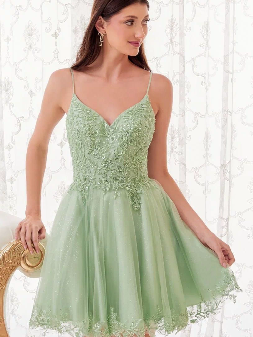 A woman wears the Short Homecoming Dress CD0213 with embroidery, featuring light green lace and tulle, sleeveless and short, as she stands beside a chair against a sheer, patterned curtain.