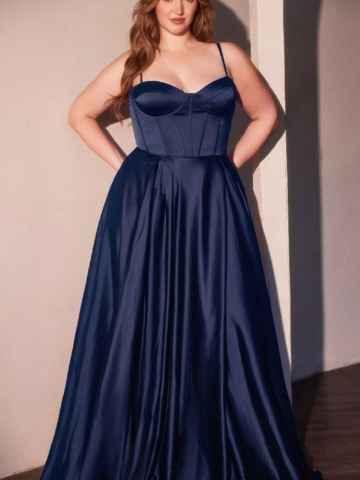 A woman stands indoors wearing the Ladivine CD337C Plus-Size Long A-Line Prom Dress, a navy blue satin gown with thin straps and a fitted bodice, her hands resting on her hips.