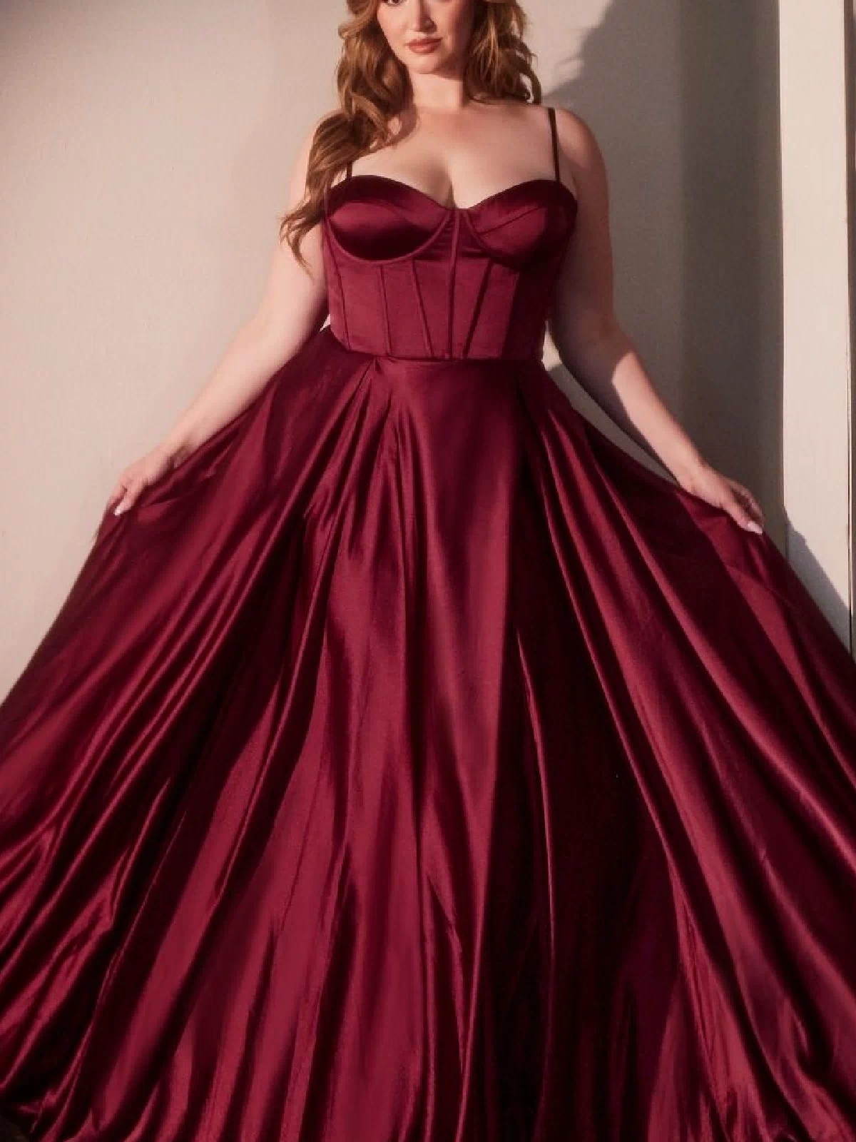 A woman wears the Ladivine CD337C Plus-Size Long A-Line Prom Dress, featuring a fitted bodice and full skirt in burgundy satin, as she stands against a pale wall holding out the sides of her gown.