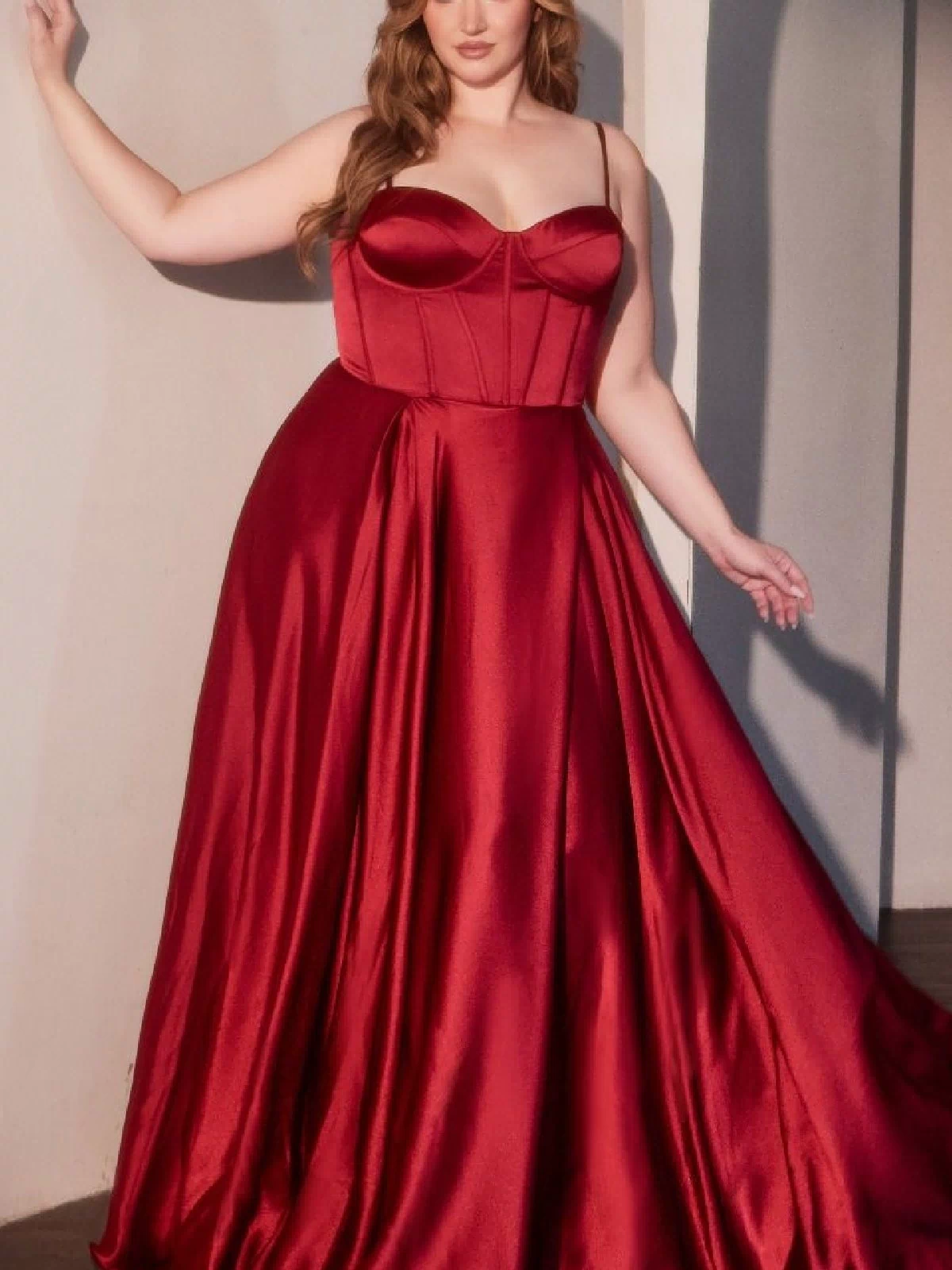 A woman poses indoors in the Ladivine CD337C, a plus-size long A-line prom dress featuring a sleeveless, fitted bodice and full skirt in striking red satin.