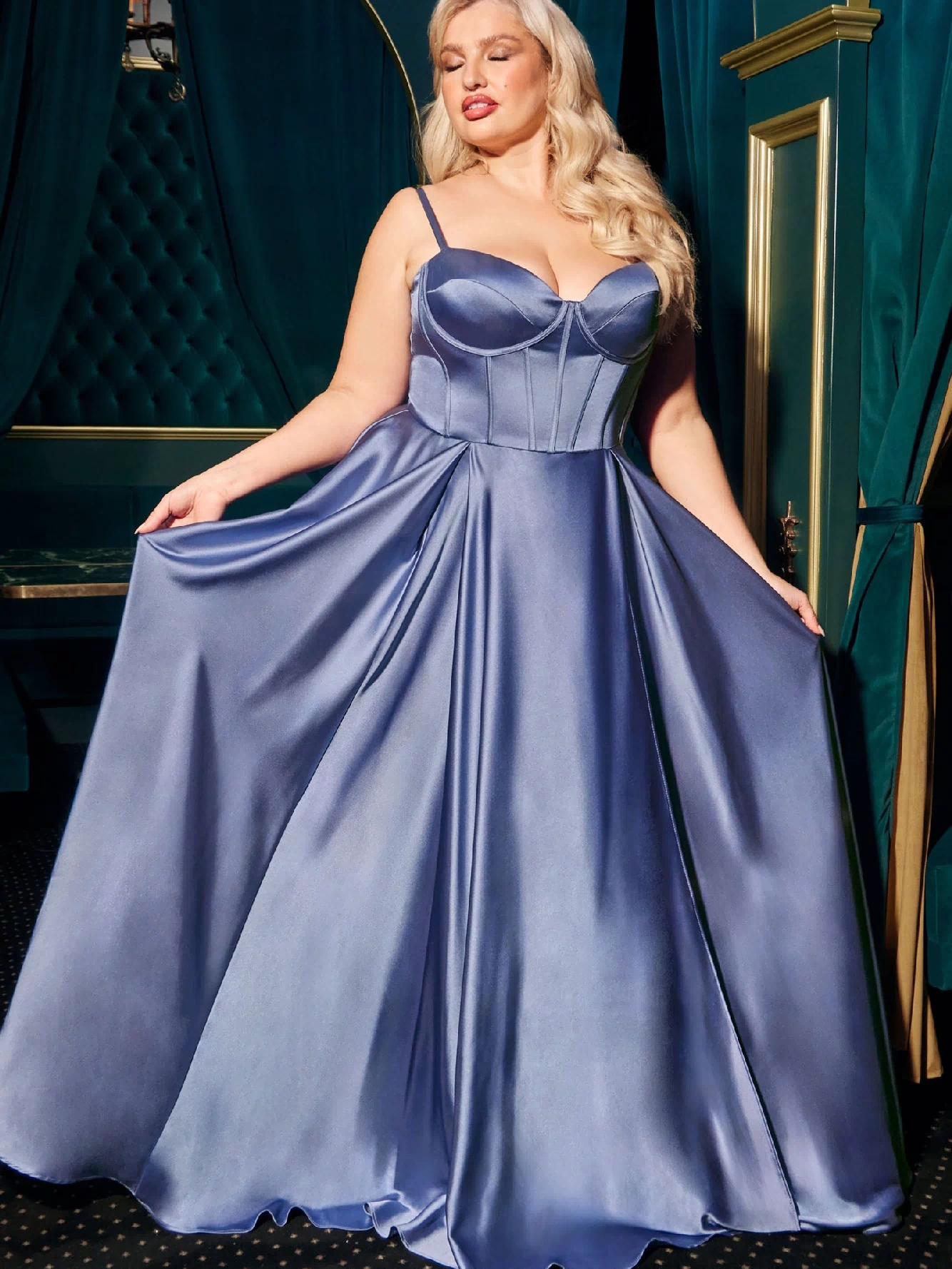 A woman in a shiny, floor-length Ladivine CD337C plus-size long A-line prom dress with a fitted bodice and thin straps holds out her gown in an elegant room with green and gold decor.