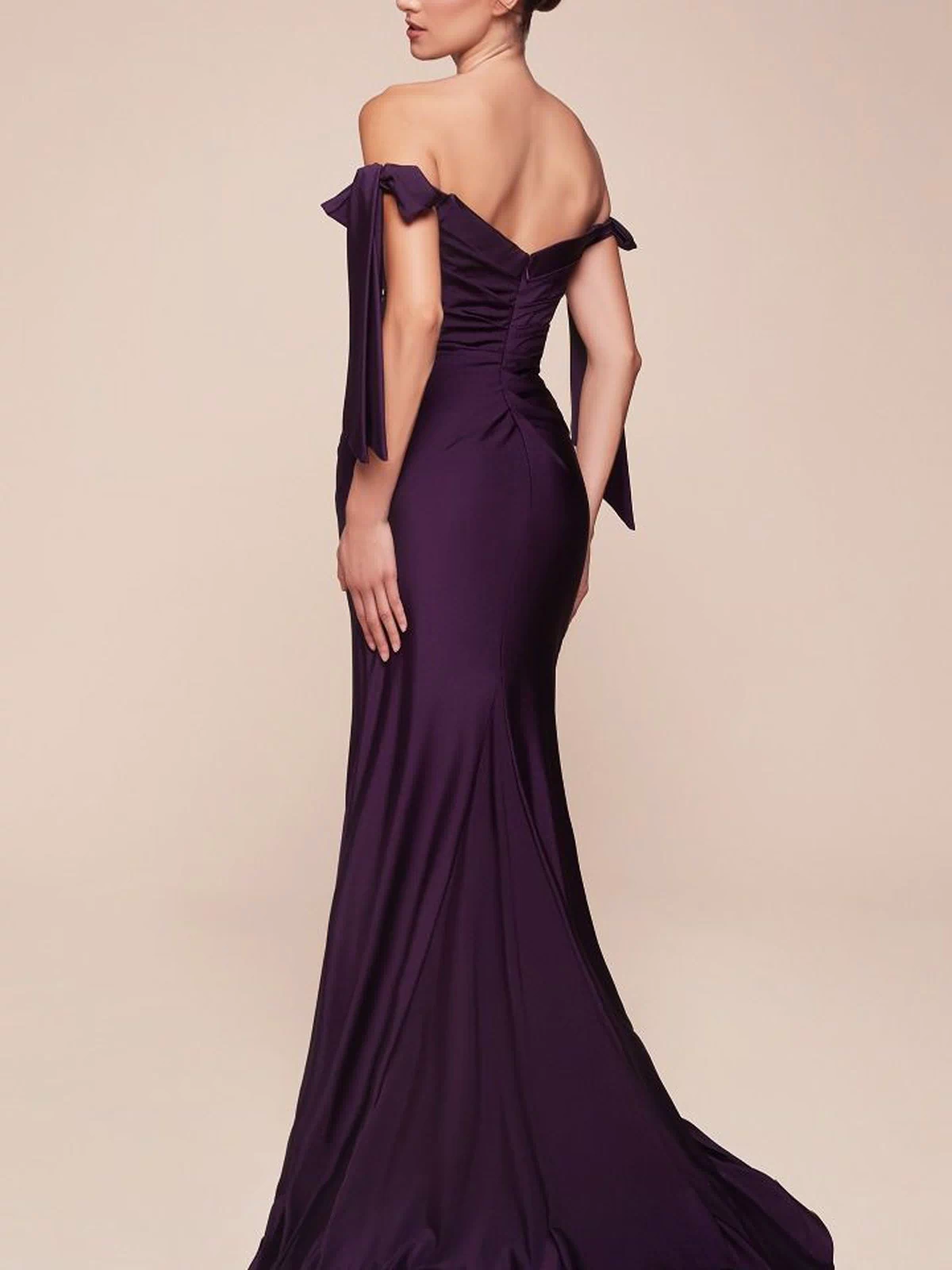 A woman in the Off-Shoulder Ruched Prom Dress: Ladivine CD943 stands with her back partially turned, highlighting the dress's fitted bodice and flowing stretch jersey floor-length design.
