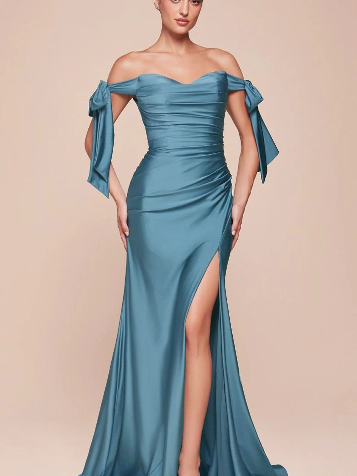 A woman stands against a plain beige background wearing the Off-Shoulder Ruched Prom Dress: Ladivine CD943 in blue, featuring off-the-shoulder sleeves, a side slit, and bow details.