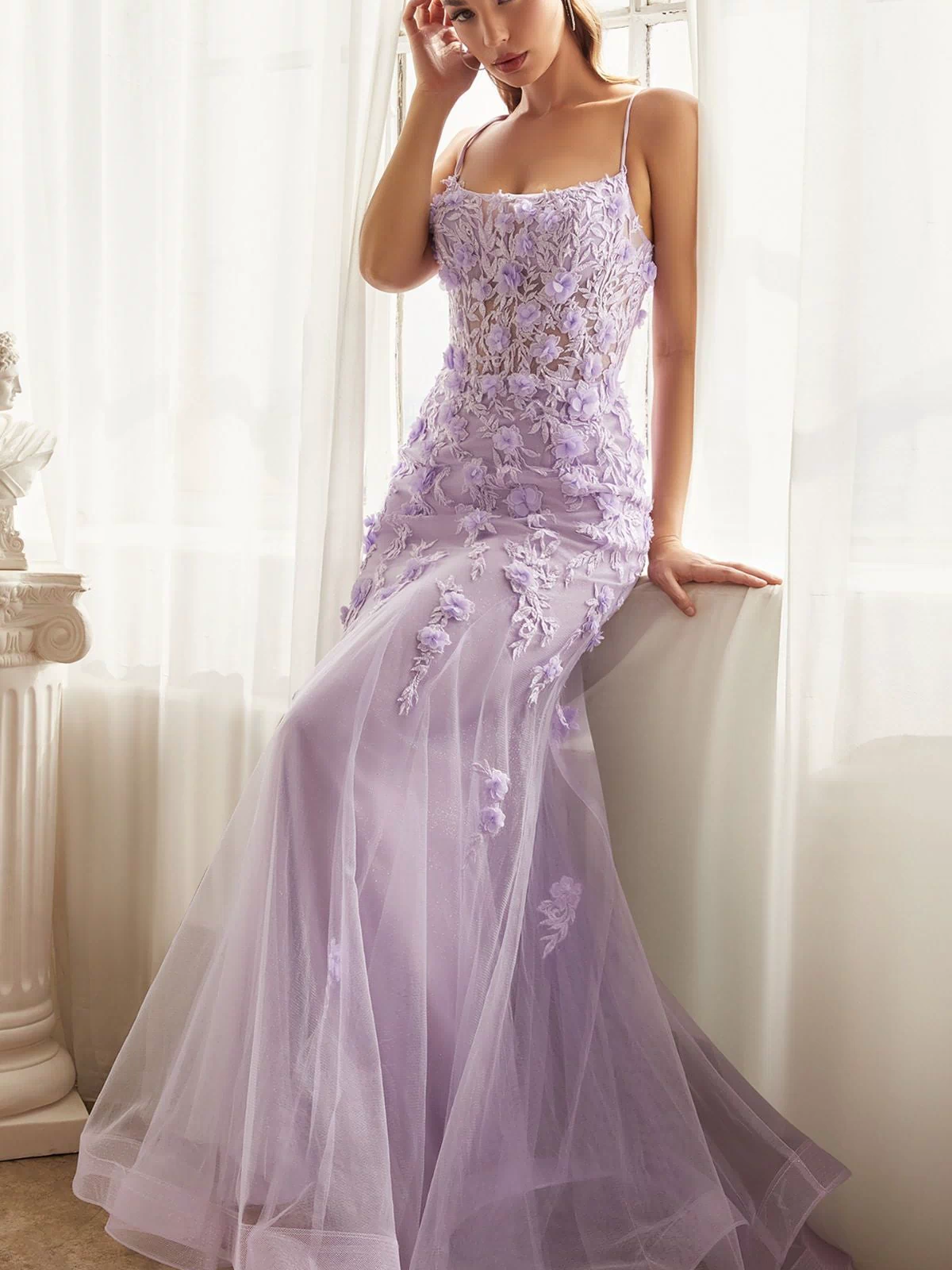 A woman stands indoors by a window with white curtains, wearing the Floral-Applique Long Mermaid Prom Dress CD995 in lavender, featuring sheer fabric and floral embroidery.