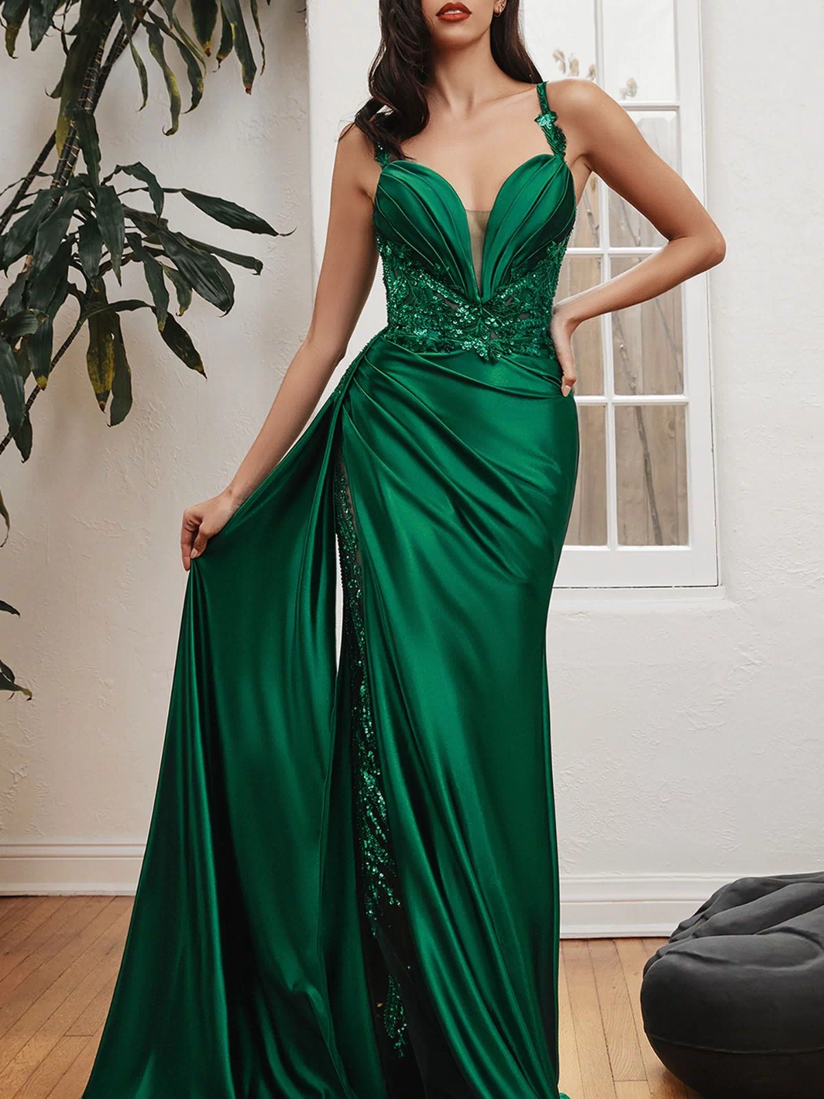 A woman stands indoors in the Long Satin Formal Dress with Side Sash CDS417, an emerald green floor-length gown featuring a deep neckline, lace details, a high slit, and holding up one side of the elegant satin dress.