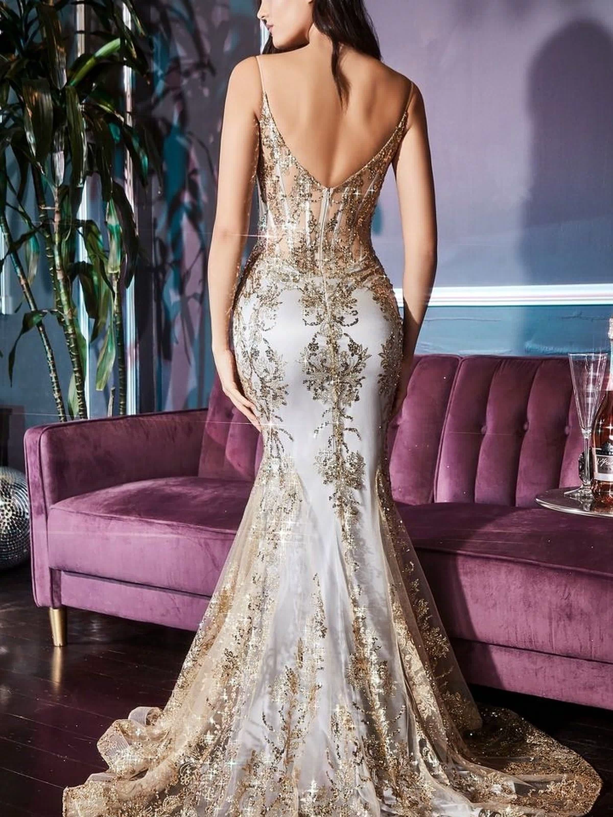 Wearing the Glitter-Print Long V-Neck Mermaid Prom Dress J810A, a woman with a corset-style bodice and intricate gold and white glitter details stands before a purple sofa in a stylish room.