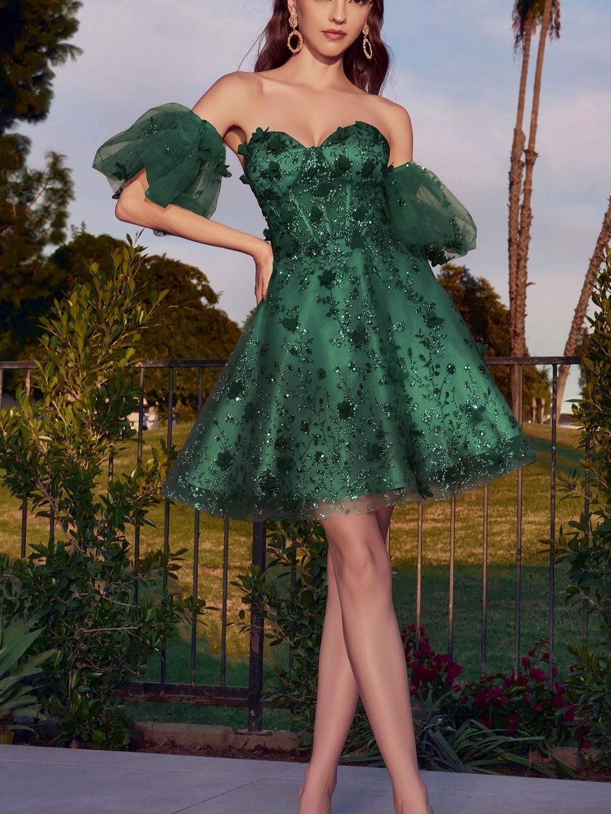 A woman stands outdoors on a patio wearing the Puff-Sleeve Short Strapless Homecoming Dress KV1089, featuring green floral embellishments and tulle puff sleeves.