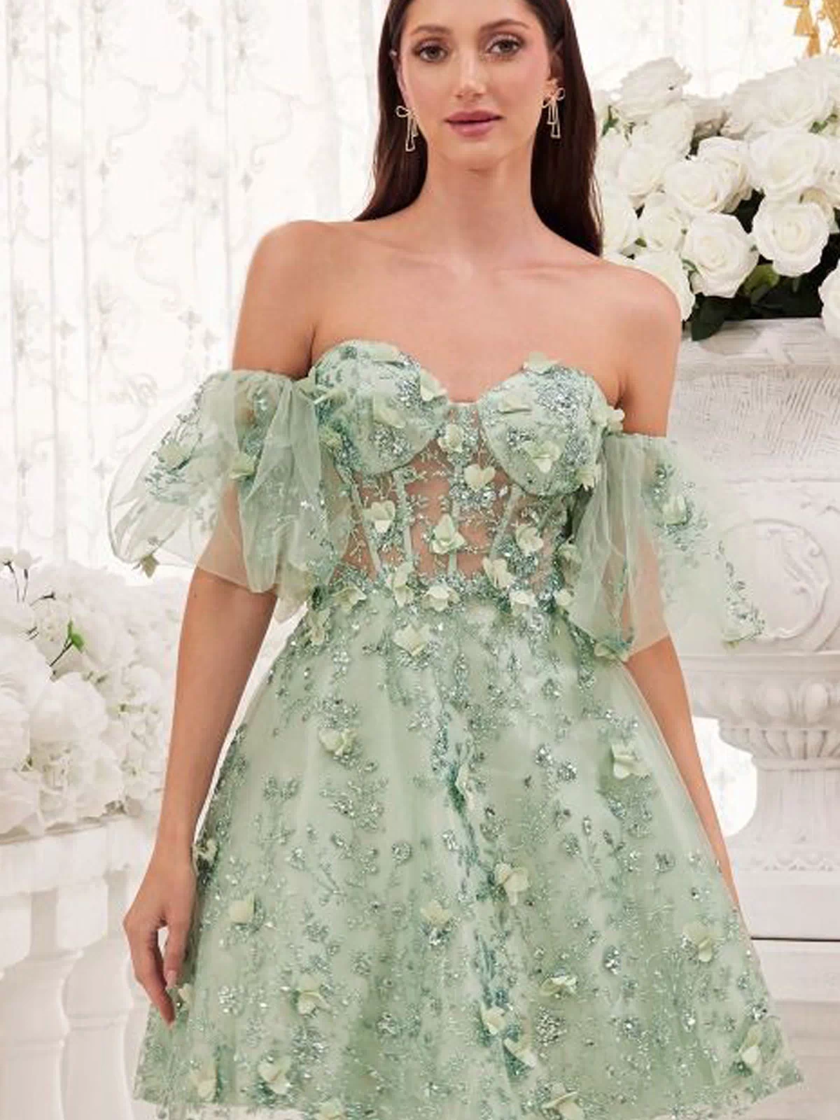 A woman wearing the Puff-Sleeve Short Strapless Homecoming Dress KV1089, a pastel green A-line party dress with floral embellishments and off-the-shoulder tulle sleeves, stands indoors near white flowers and ornate decor.