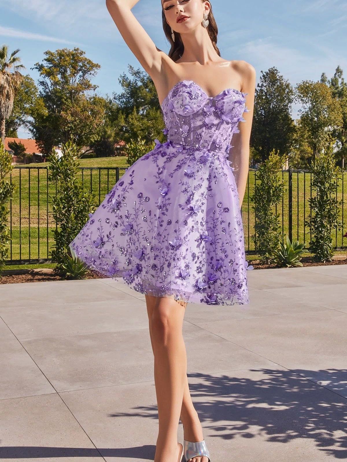 A woman poses outdoors in the Puff-Sleeve Short Strapless Homecoming Dress KV1089 with lilac floral embellishments and silver heels, one arm raised overhead; trees and a fence form the background.