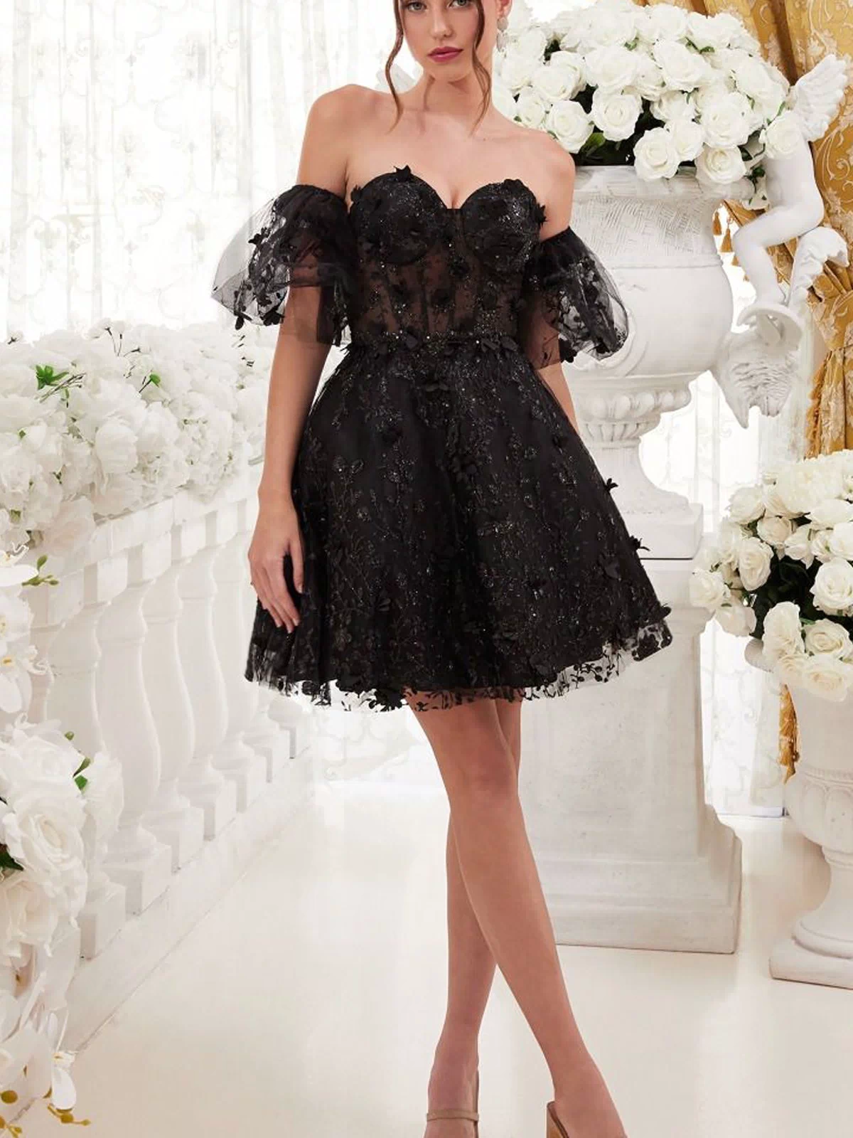 A woman stands indoors wearing the Puff-Sleeve Short Strapless Homecoming Dress KV1089 paired with nude heels. White flowers and gold curtains accent the background.