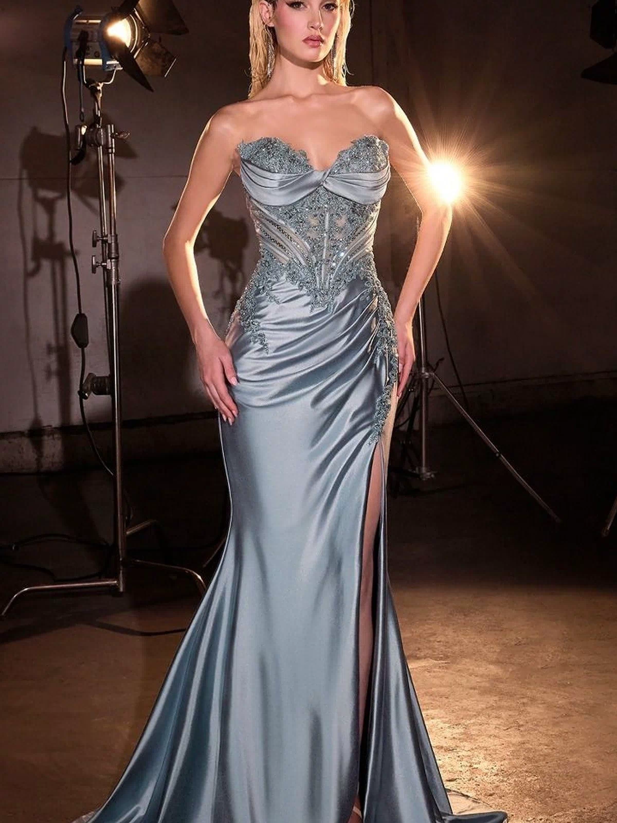 A woman wears the Strapless Long Prom Dress: Ladivine KV1132, a light blue satin gown with a beaded corset bodice, intricate detailing, and high leg slit, posing in front of studio lights.