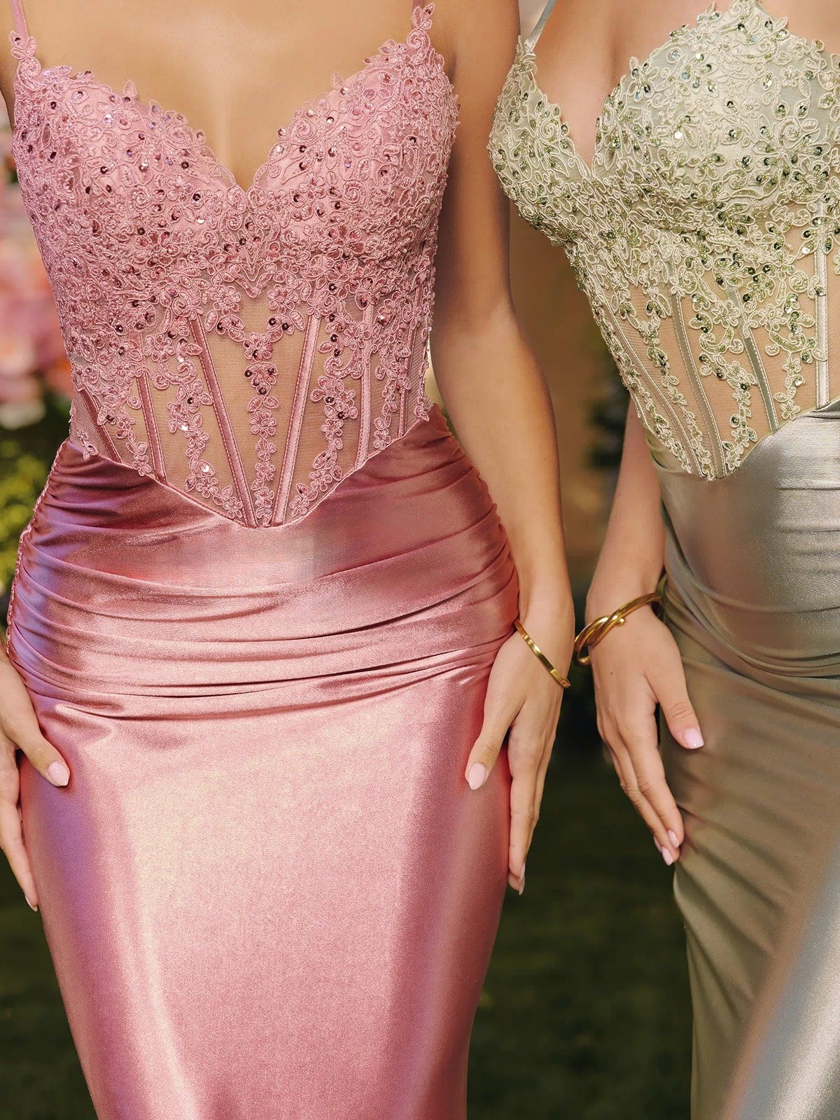Two women wear the Amoris T819 Long Prom Dress: one in pink and one in silver metallic stretch satin, both with fitted embellished corset bodices, lace, and sequin details. Their heads are not visible in the image.