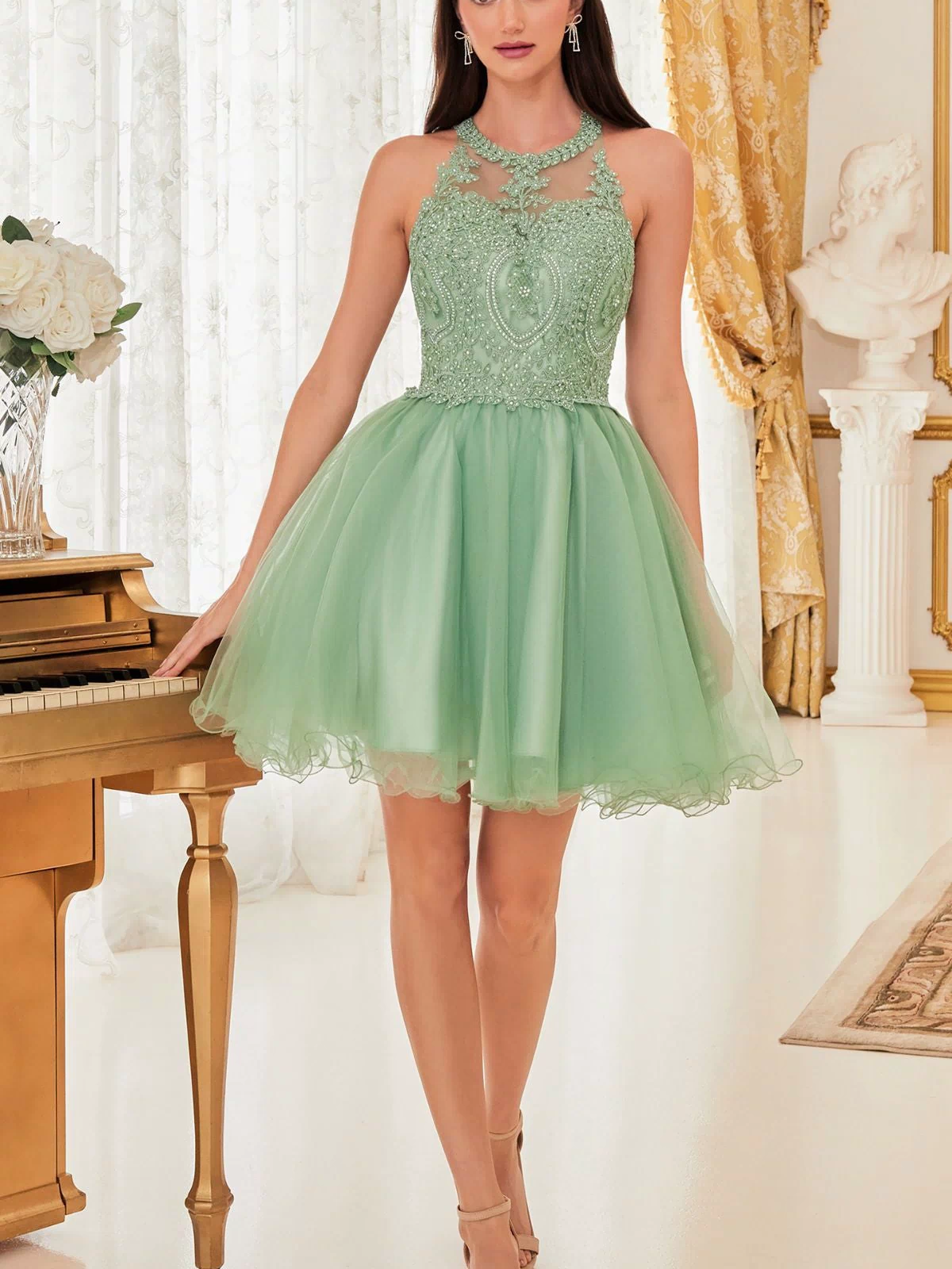 A woman stands indoors near a gold piano, wearing the High-Neck Short Babydoll Homecoming Dress UJ0119—sleeveless, mint green, knee-length with an embroidered-lace bodice and tulle skirt—surrounded by elegant white and gold decor.