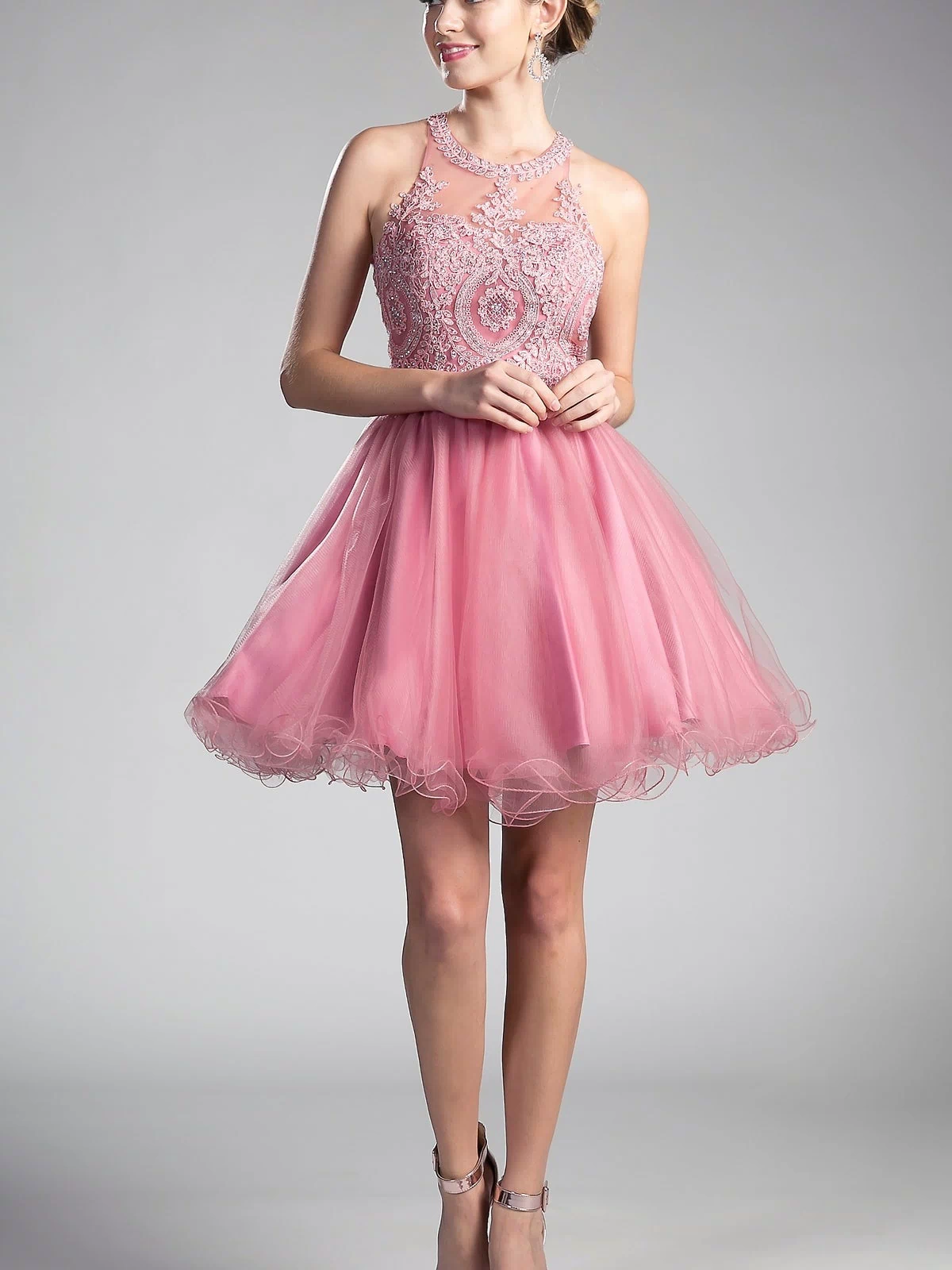 A woman in the High-Neck Short Babydoll Homecoming Dress UJ0119, featuring a sleeveless pink tulle skirt and beaded bodice, pairs it with silver heels while standing against a plain gray background.