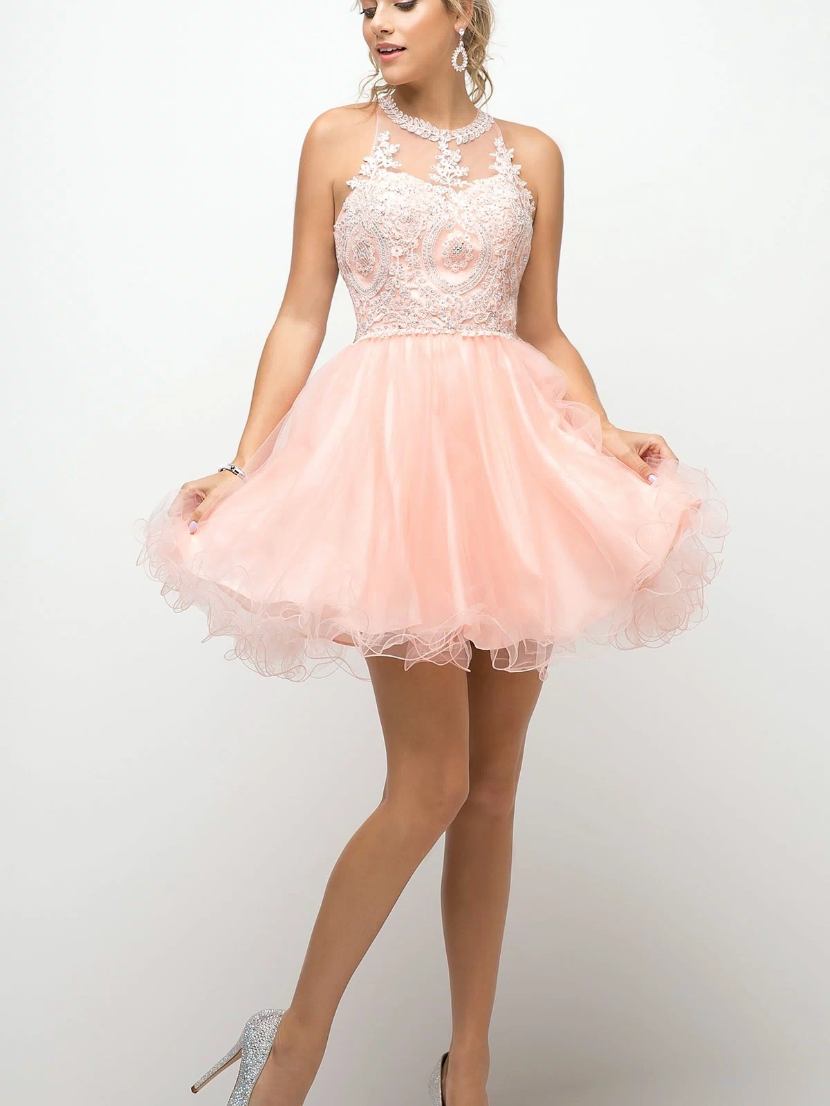 A woman in the High-Neck Short Babydoll Homecoming Dress UJ0119—a sleeveless, light pink tulle dress with an embroidered-lace bodice and silver heels—stands against a plain white background.