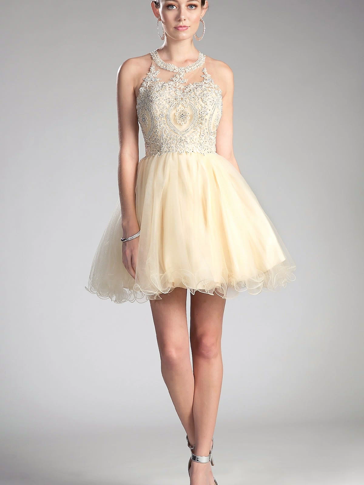 A young woman stands in the High-Neck Short Babydoll Homecoming Dress UJ0119 with an embroidered-lace bodice and tulle skirt, silver heels, and hoop earrings—a stylish, affordable cocktail dress for any occasion.