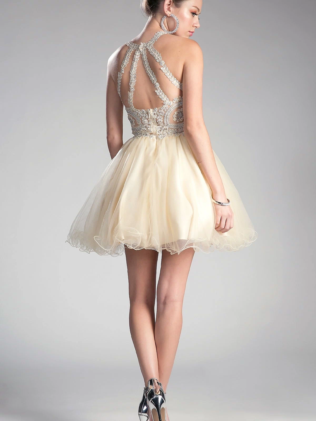 A woman stands against a neutral background wearing the affordable High-Neck Short Babydoll Homecoming Dress UJ0119, featuring an embroidered lace bodice, sleeveless and backless design, full cream skirt, and silver high heels.