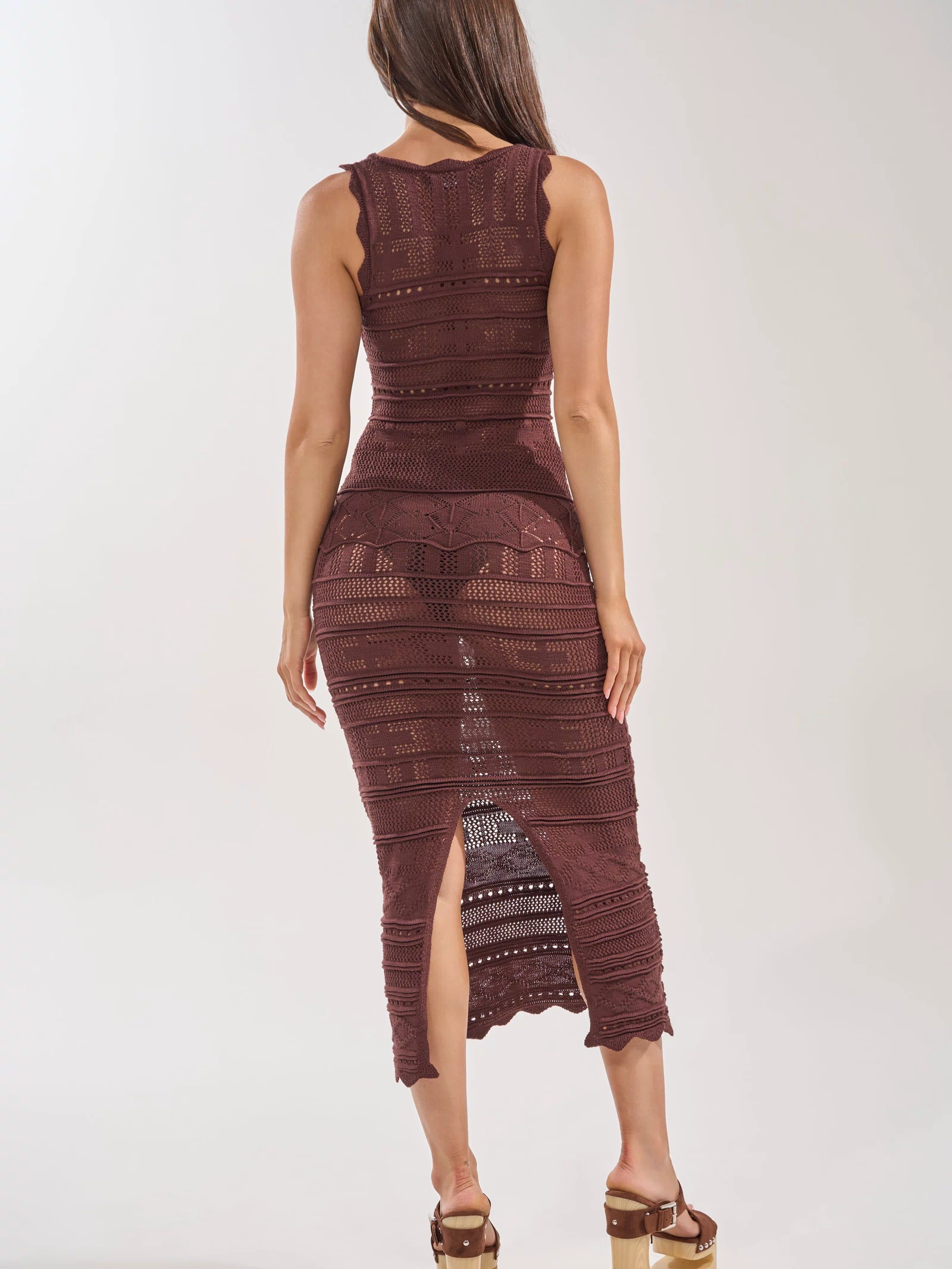 A woman with long brown hair wears the LONG WALKS ON THE BEACH CROCHET MAXI DRESS, a sleeveless maroon maxi with intricate crochet details, perfect for vacation. She faces away, highlighting the knit pattern and beige platform sandals.
