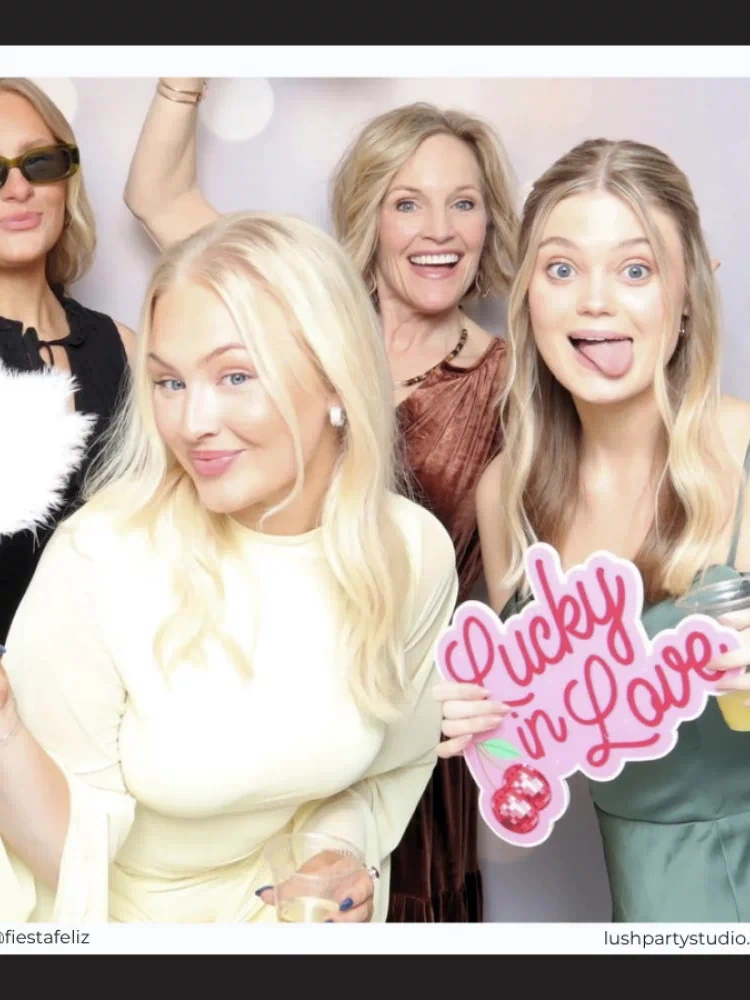 Four women posing together with a 'Lucky in Love' sign in a photo booth setting.