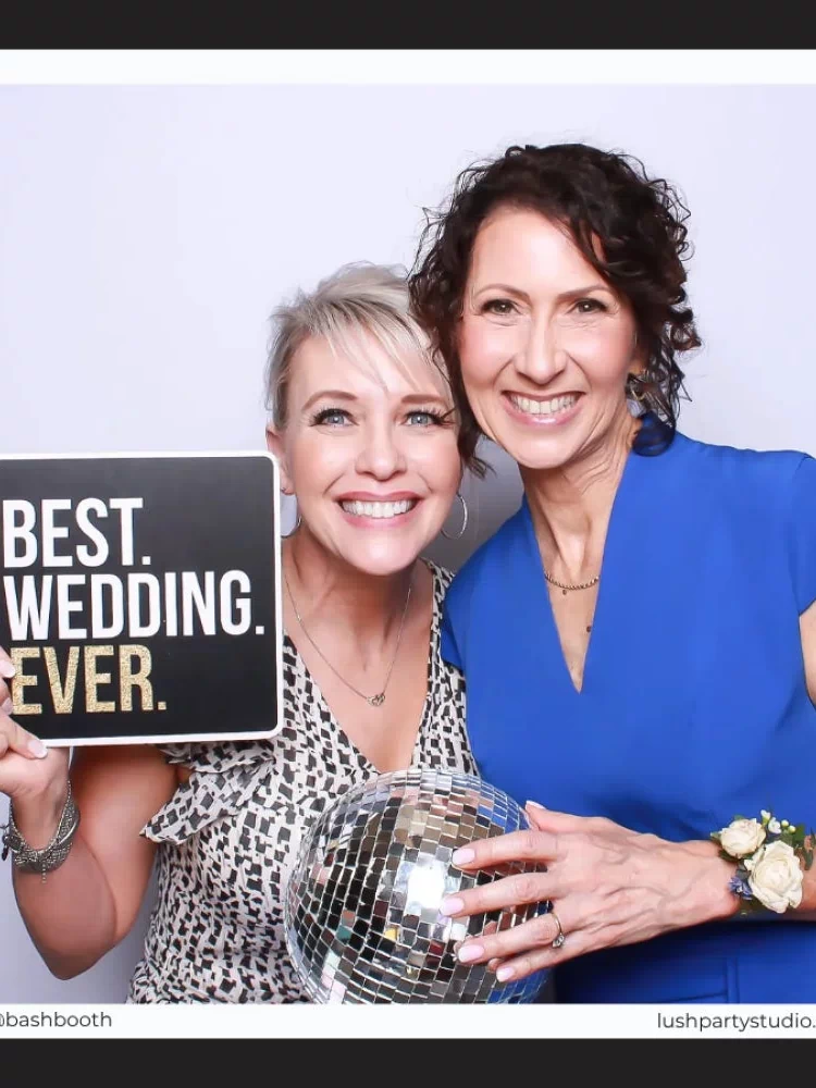Two women holding a disco ball and a photo booth prop sign that reads 'Best Wedding Ever' by Lush Party Studio