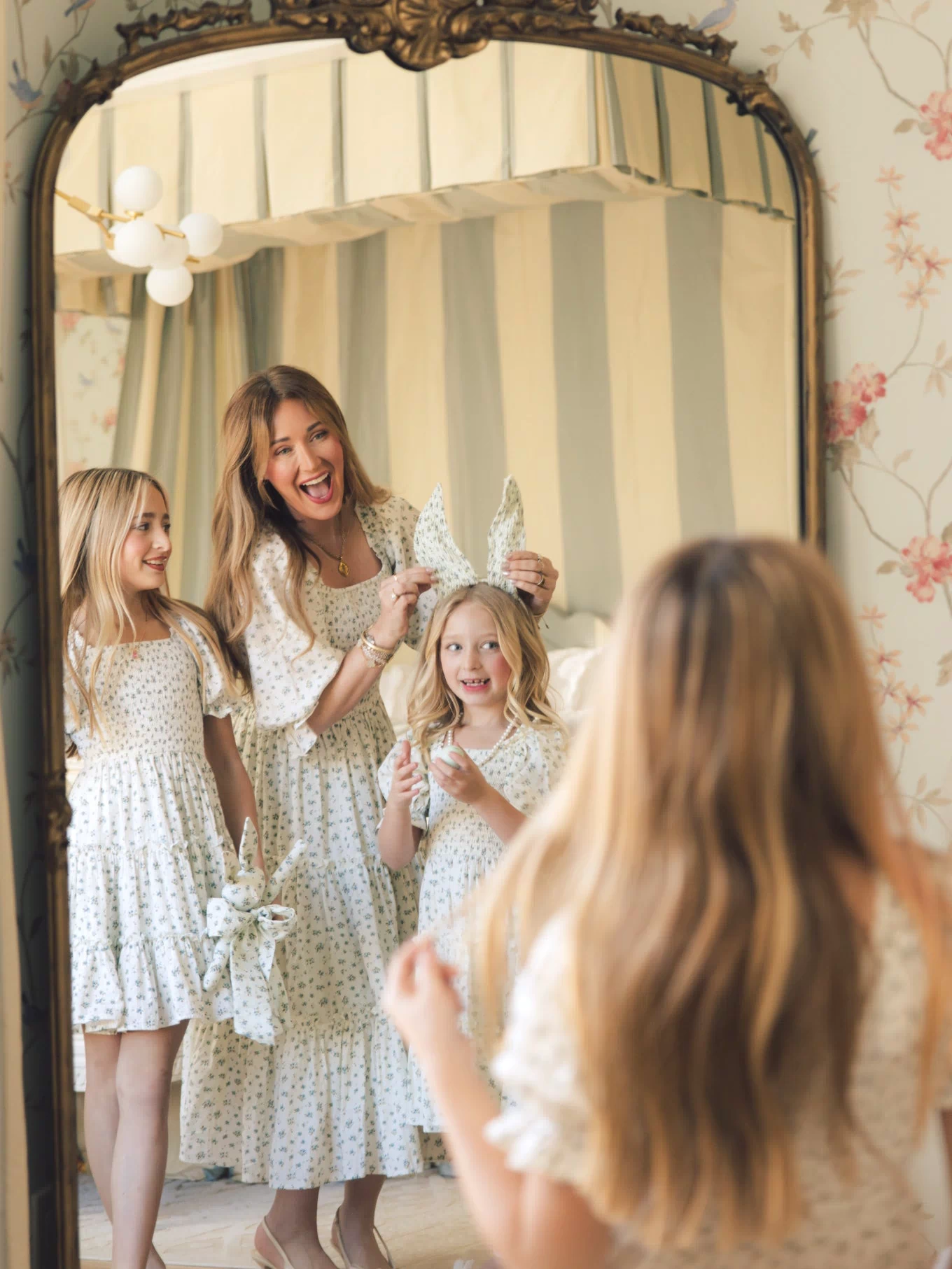 A woman wears the Ivy City Co Madeline Dress in Blue, smiling with two young girls in matching outfits by an antique mirror in a vintage-style room, holding playful bunny ear headbands.