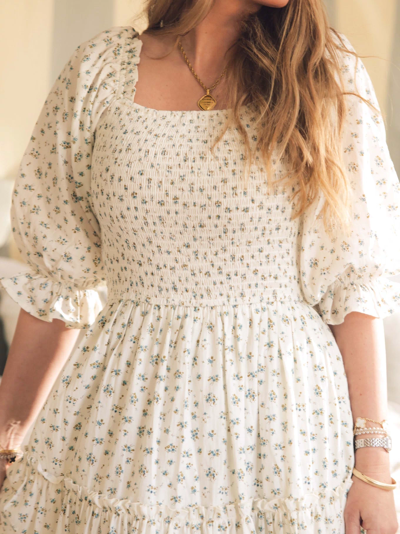 A woman models the Ivy City Co Madeline Dress in Blue, a maternity and nursing-friendly midi with a smocked bodice and puffed sleeves, styled with gold jewelry. Soft lighting showcases her long hair and the dress’s elegant floral design.