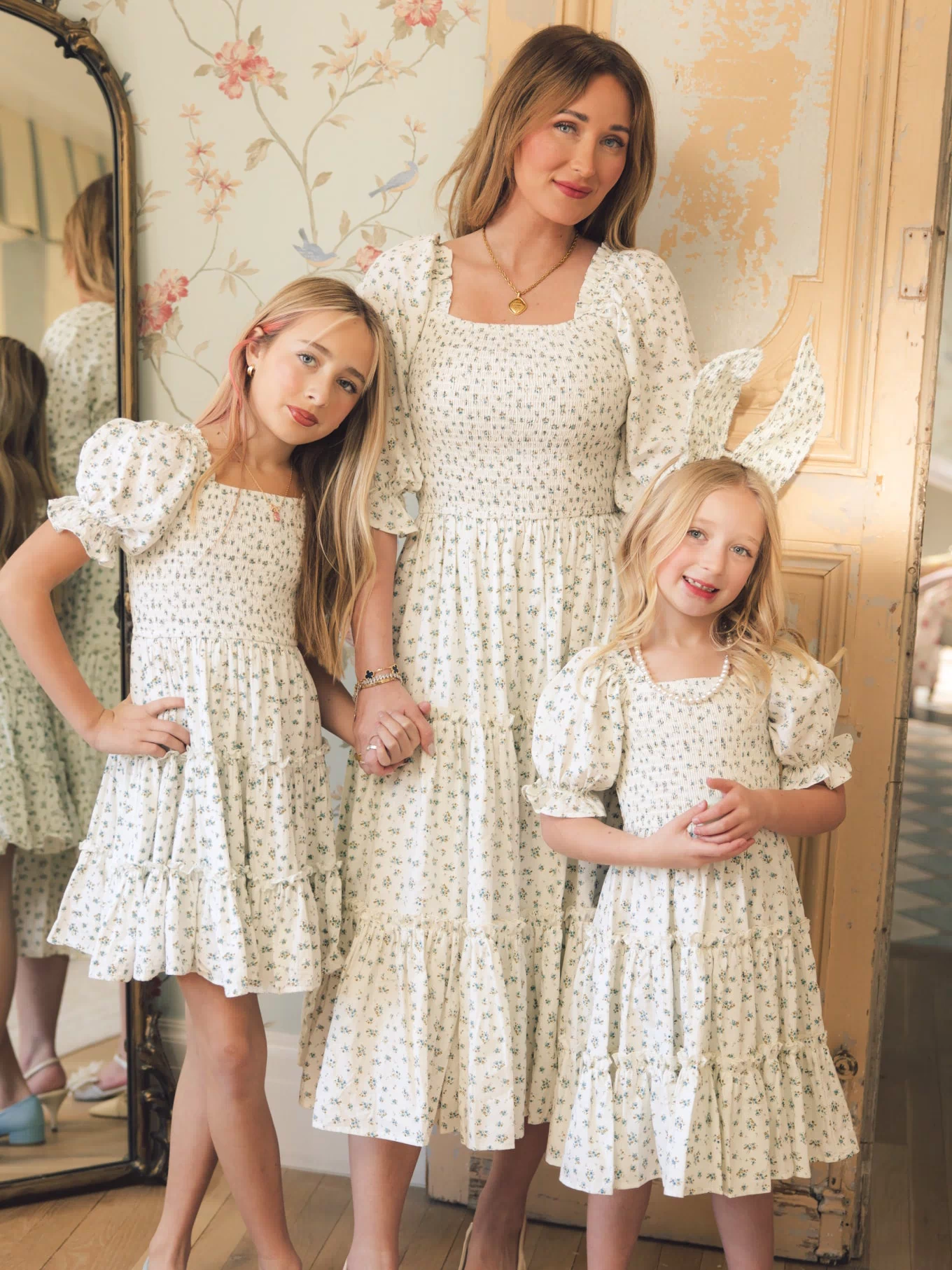 A woman and two young girls smile indoors, holding hands and wearing matching Ivy City Co Madeline Dress in Blue with small floral prints and a smocked bodice, posing in front of a mirror and vintage-style door.