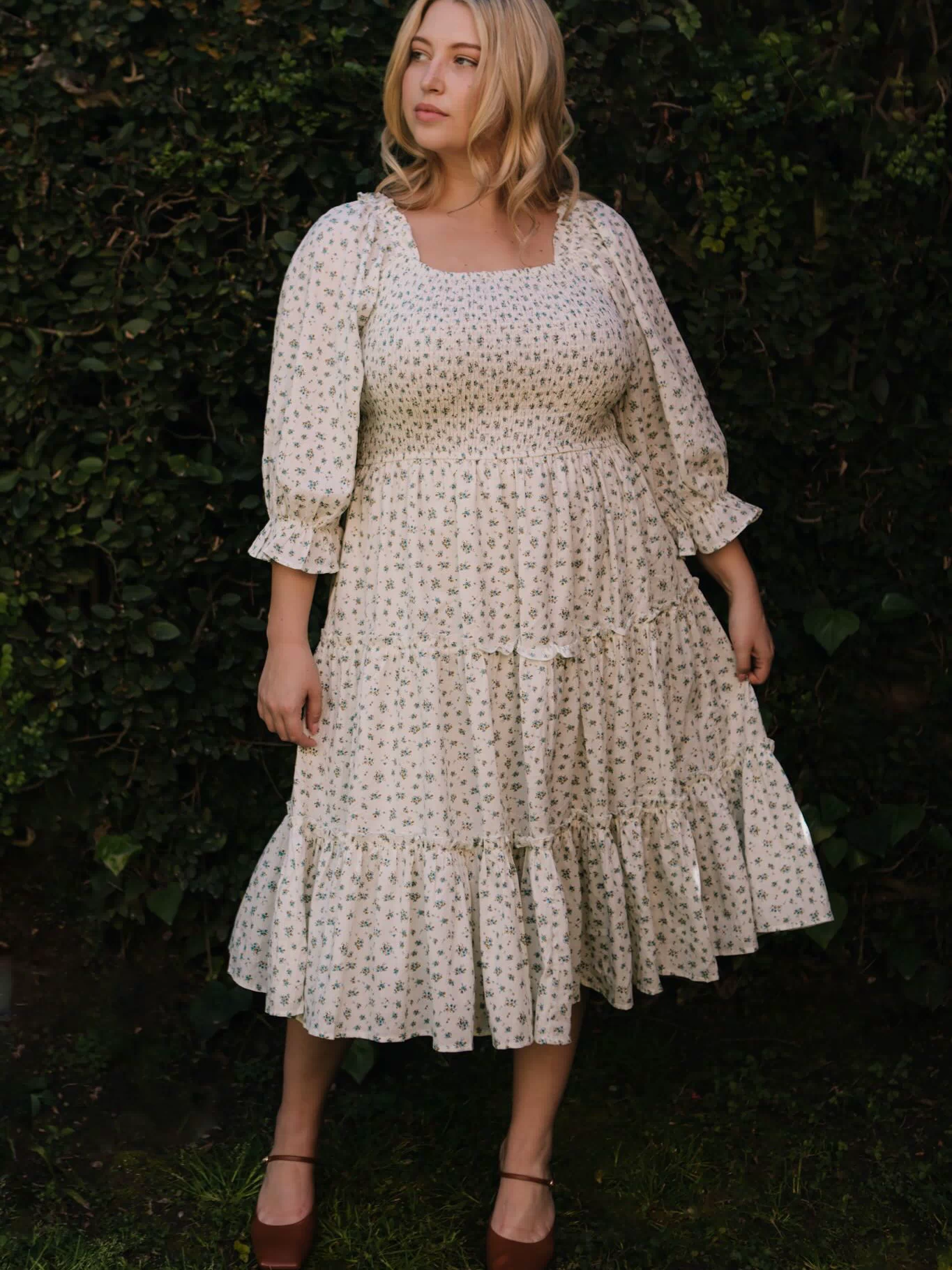 A blonde woman stands outdoors before green bushes, wearing the Ivy City Co Madeline Dress in Blue—a floral, smocked midi that's maternity and nursing friendly—paired with brown platform shoes. She gazes calmly to the side.